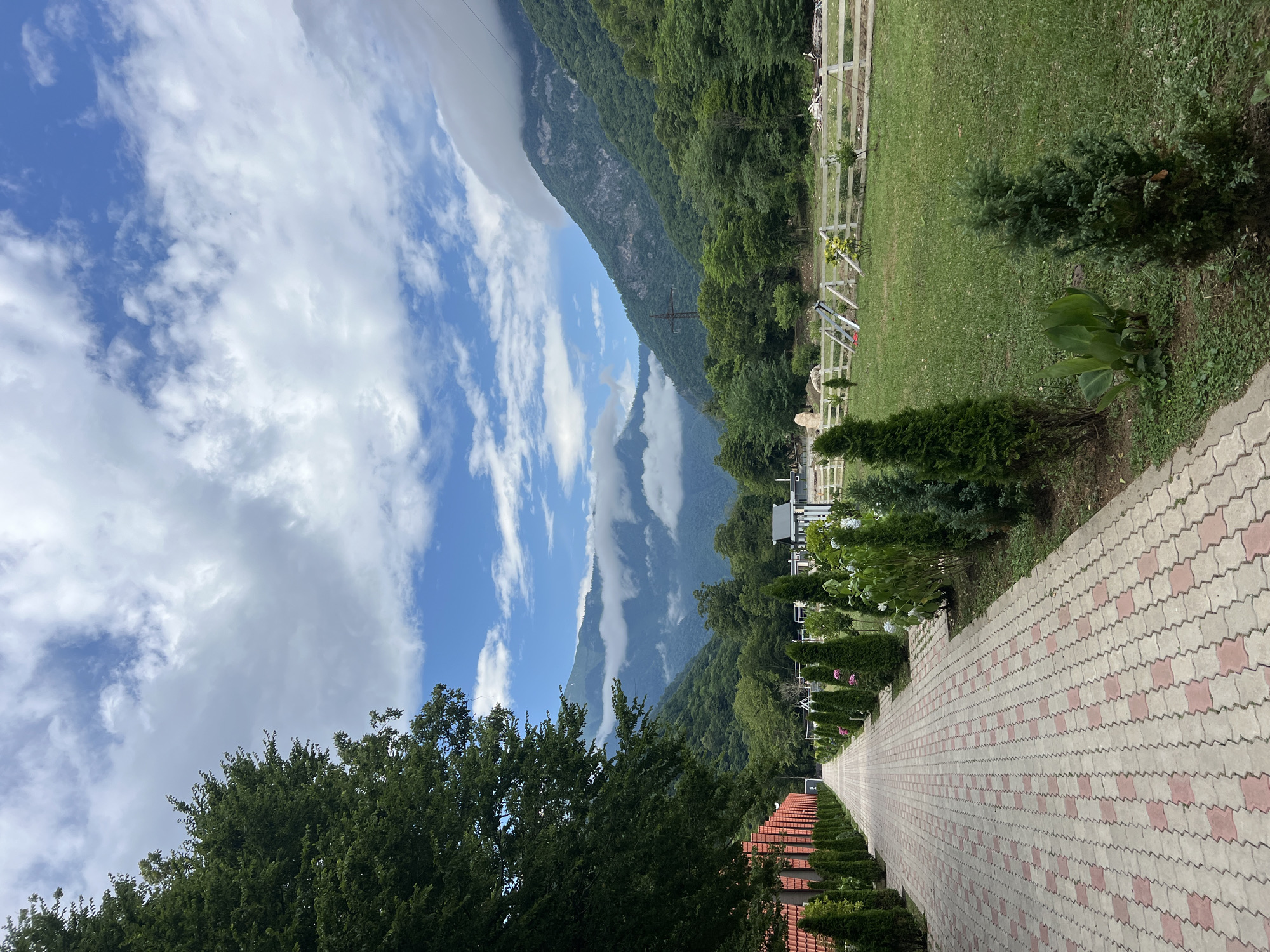 The image depicts a paved walkway lined with manicured hedges and trees, leading towards a backdrop of lush green mountains partially obscured by clouds. To the left of the walkway, a red-tiled wall runs along the path. To the right of the walkway, a grassy area is enclosed by a white wooden fence, with some farm structures visible in the distance. The sky is partly cloudy with patches of blue. The scene appears to be a tranquil outdoor setting, possibly a park or a rural resort.