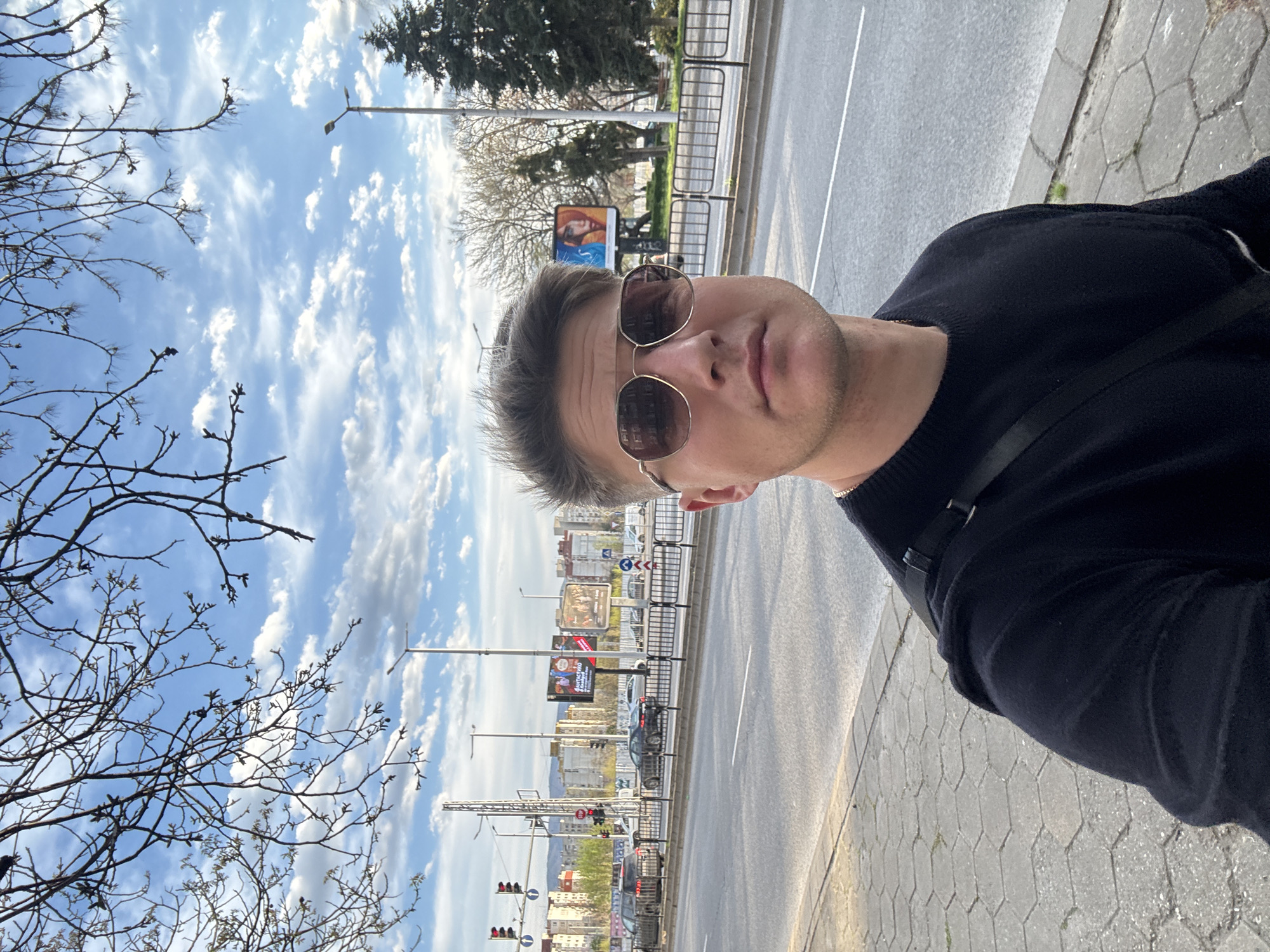 A man wearing sunglasses and a dark sweater stands on a sidewalk next to a street in Plovdiv, Bulgaria. The street has multiple lanes and is lined with billboards and lamp posts. Buildings are visible in the background. A tree with bare branches is visible to the left. The sky is blue with scattered clouds.
