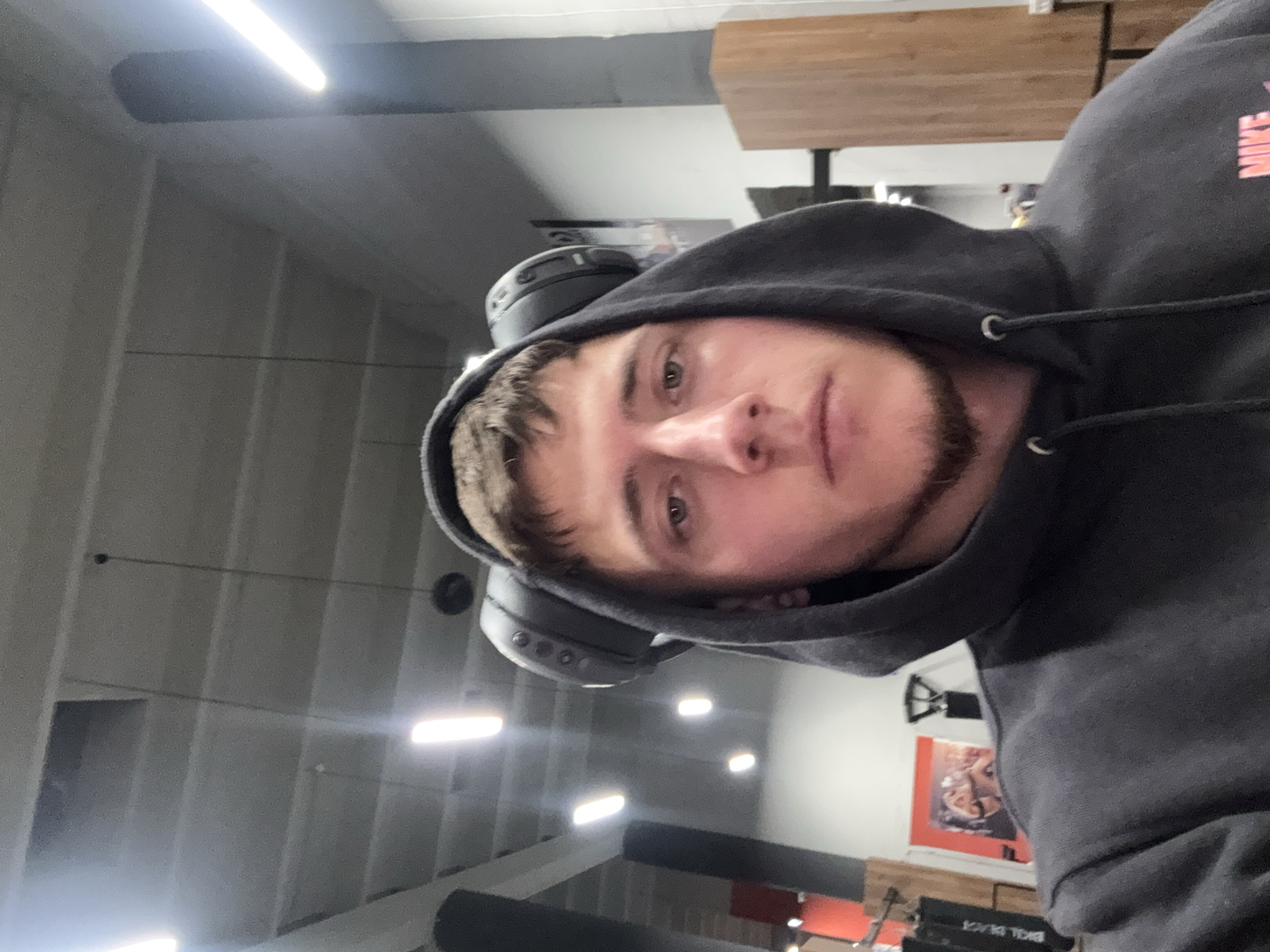 The image is a head-and-shoulder self-portrait of a male subject in an indoor, commercial setting, identified as a gym. The subject, appearing to be in his early twenties, has short brown hair, light eyes, and a short beard. He wears a dark grey hooded sweatshirt with the hood up, partially revealing a "NIKE" logo in pink lettering on the lower right chest area. Black over-ear headphones rest atop his head, obscured by the hood. His expression is neutral as he gazes directly into the camera.
The background features an industrial-style ceiling with exposed beams or panels, illuminated by multiple rectangular LED strip lights and containing circular dark fixtures. Dark structural columns are visible on both sides of the frame. To the left of the subject, gym equipment is partially visible, including a red apparatus, a black vertical structure, and a framed poster depicting two figures, possibly engaged in a sport, against an orange background. To the right, a tall, light-colored wooden paneled structure is present. The environment is consistent with a modern fitness facility in Sofia, Bulgaria. The specific location within the city cannot be determined from the visual information provided.