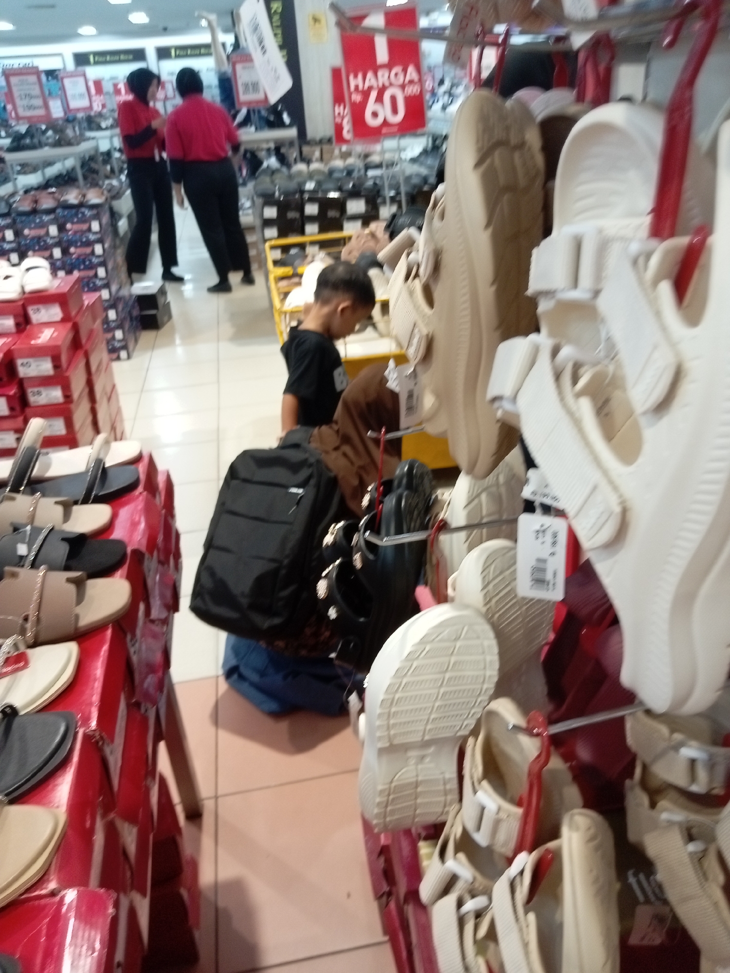 The image depicts the interior of a shoe store in Batam, Indonesia, with a focus on the display of various footwear. In the foreground, a collection of women's sandals is prominently featured, arranged on tiered red displays. These sandals are in various colors including beige, black, and white, with some featuring decorative elements like chains and buckles. To the right, a display of white platform sandals or clogs is visible, with a tag attached to one pair.
In the mid-ground, a young boy is seen wearing a black t-shirt, looking at the footwear displays. He is partially obscured by a black backpack that he is carrying. Behind him, two store employees wearing red shirts and black pants are conversing. The store environment is characterized by shelves stocked with shoe boxes in the background, indicating a retail setting. Additionally, price tags and promotional signs, such as one reading "HARGA 60", are visible, suggesting ongoing sales or pricing information. The floor is tiled, and the overall lighting suggests an indoor commercial space.