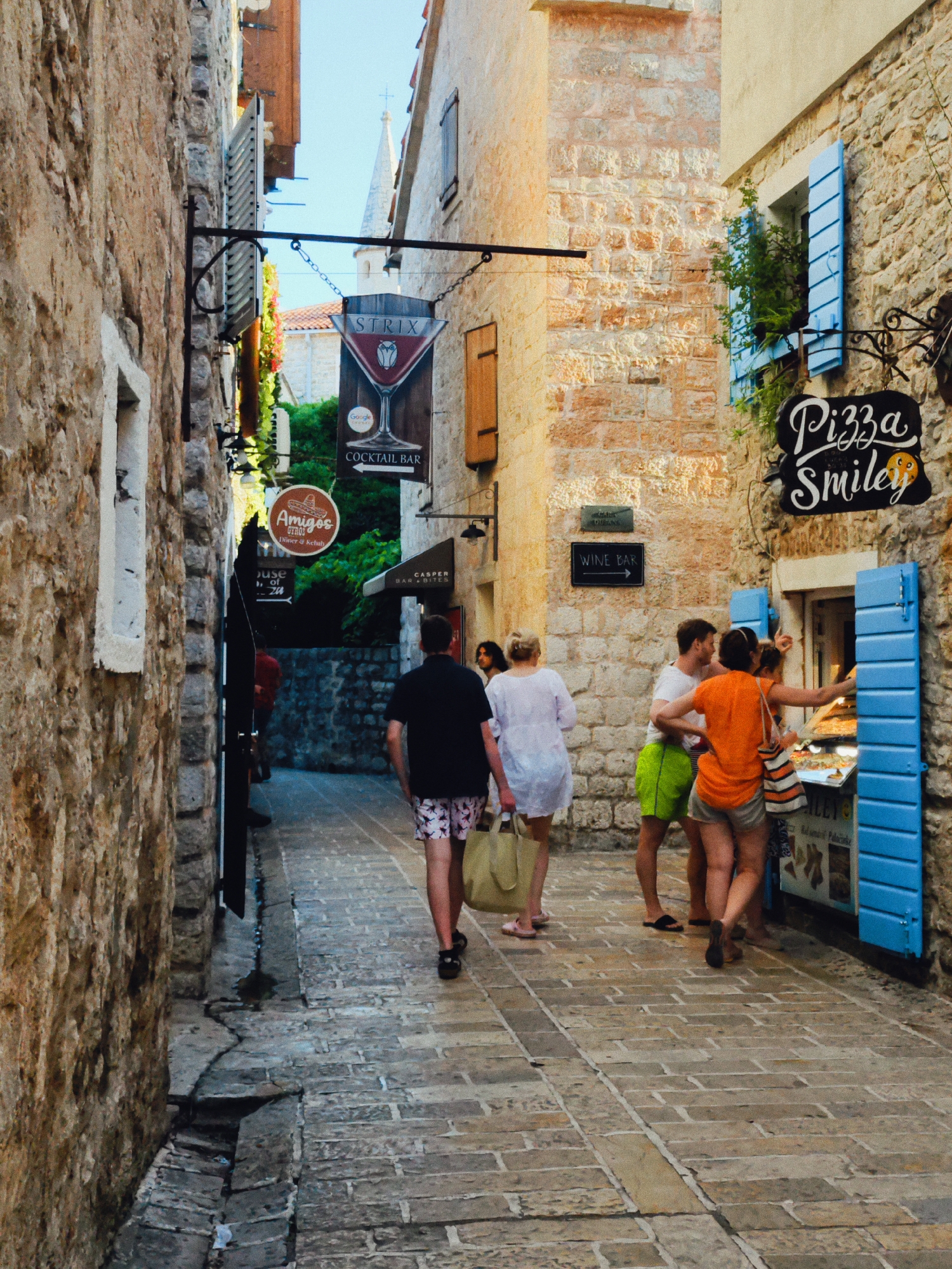 This photograph captures a lively street scene in Budva, Montenegro, bathed in what appears to be bright daylight, possibly late morning or afternoon. The setting is a narrow, cobblestone alleyway lined with old stone buildings adorned with wooden shutters and signs for various establishments.
In the foreground and midground, several people are casually strolling and interacting. On the left, a man in a dark t-shirt and patterned shorts walks away from the camera, carrying a large tote bag. Further down the alley, a woman in a white dress with blonde hair also walks away, holding a similar bag. Towards the right of the frame, a group of four people are gathered near a window, possibly a food stand or shop, where a woman in an orange top is reaching towards the counter. A man in a white t-shirt and bright green shorts stands next to her, while another couple is partially obscured.
Various signs are visible, indicating the commercial nature of the street. One sign for "STRIX COCKTAIL BAR" hangs prominently, featuring a martini glass graphic. Another sign reads "Amigos GYROS Döner & Kebab." Further along, a sign for "CASPER BAR & BITES" is visible, and a smaller sign points towards a "WINE BAR." At the far right, a sign with "Pizza Smiley" and a smiley face graphic suggests a pizza establishment. The blue shutters of this establishment are a striking detail against the stone facade.
The architecture, the stone paving, and the narrowness of the alley suggest a historic European town. The overall mood is relaxed and leisurely, indicative of tourists enjoying a sunny day exploring the local amenities. The presence of multiple food and drink establishments hints at a popular area for both locals and visitors.