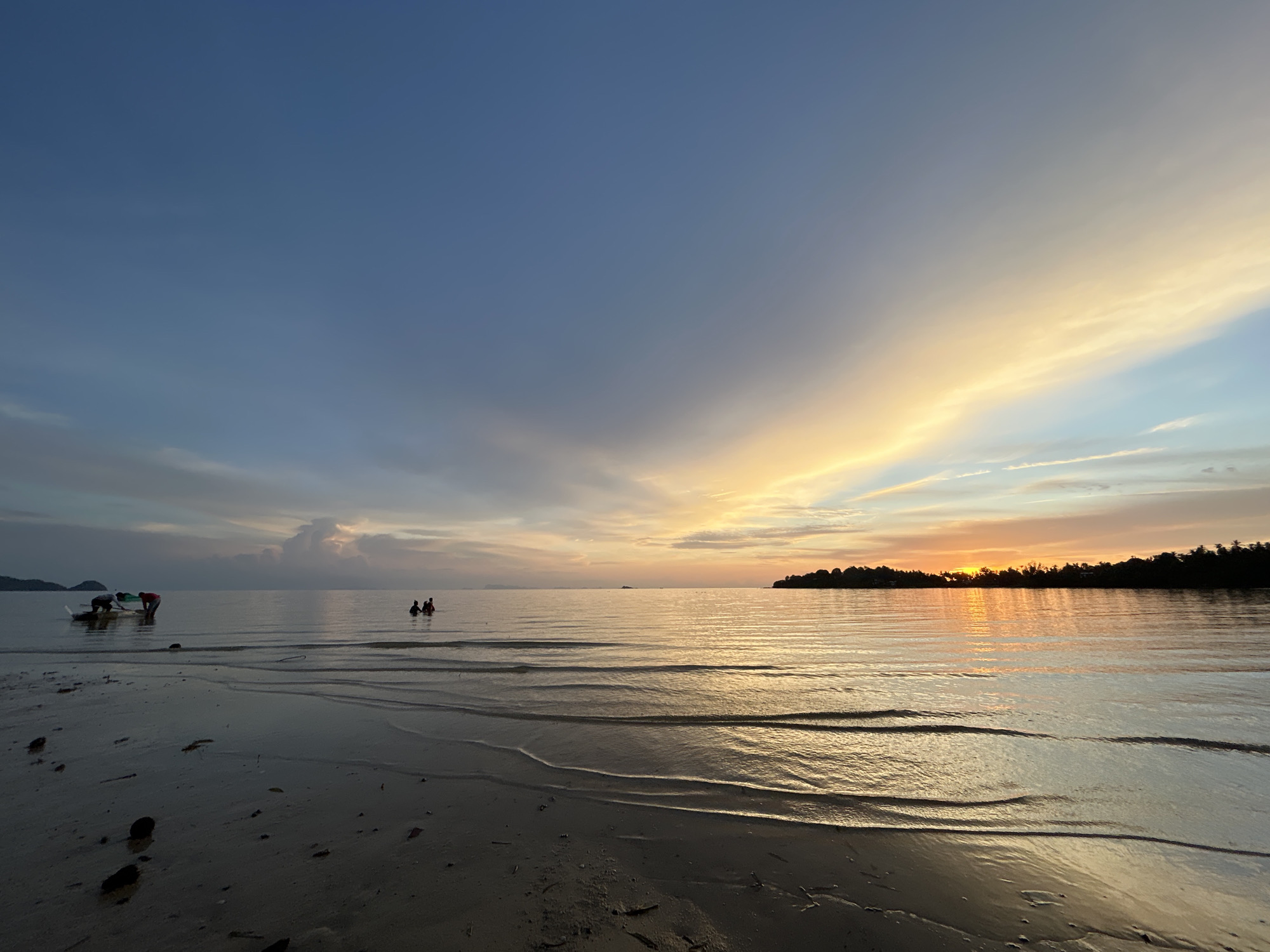 At dusk, on a tropical beach in Ko Pha-ngan District, Thailand, the sun casts a warm golden glow across the calm sea. The sky transitions from a deep blue to vibrant shades of orange and yellow, with wispy clouds catching the last light. The foreground features a sandy shore with small waves gently lapping at the wet sand, creating rippled patterns. A small boat with a few individuals is visible near the shore on the left, appearing to be preparing for or returning from an excursion. Further out in the water, a group of people stand and interact, seemingly enjoying the serene evening. The distant horizon is lined with a dark silhouette of lush trees and vegetation, marking the land. The overall atmosphere is peaceful and tranquil.