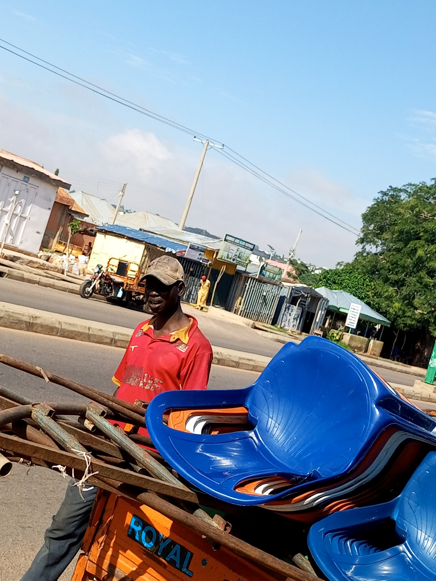 A man in a red shirt and a baseball cap stands in a busy street in Tempure, Nigeria, holding a load of blue plastic chairs and metal rods. He is likely a vendor or transporter, as the chairs are stacked on an orange trolley, with the word "ROYAL" visible on its side. The street is lined with buildings, shops, and trees, and traffic is visible in the background, including a motorcycle and a yellow truck. The sky is clear and blue, indicating a sunny day.