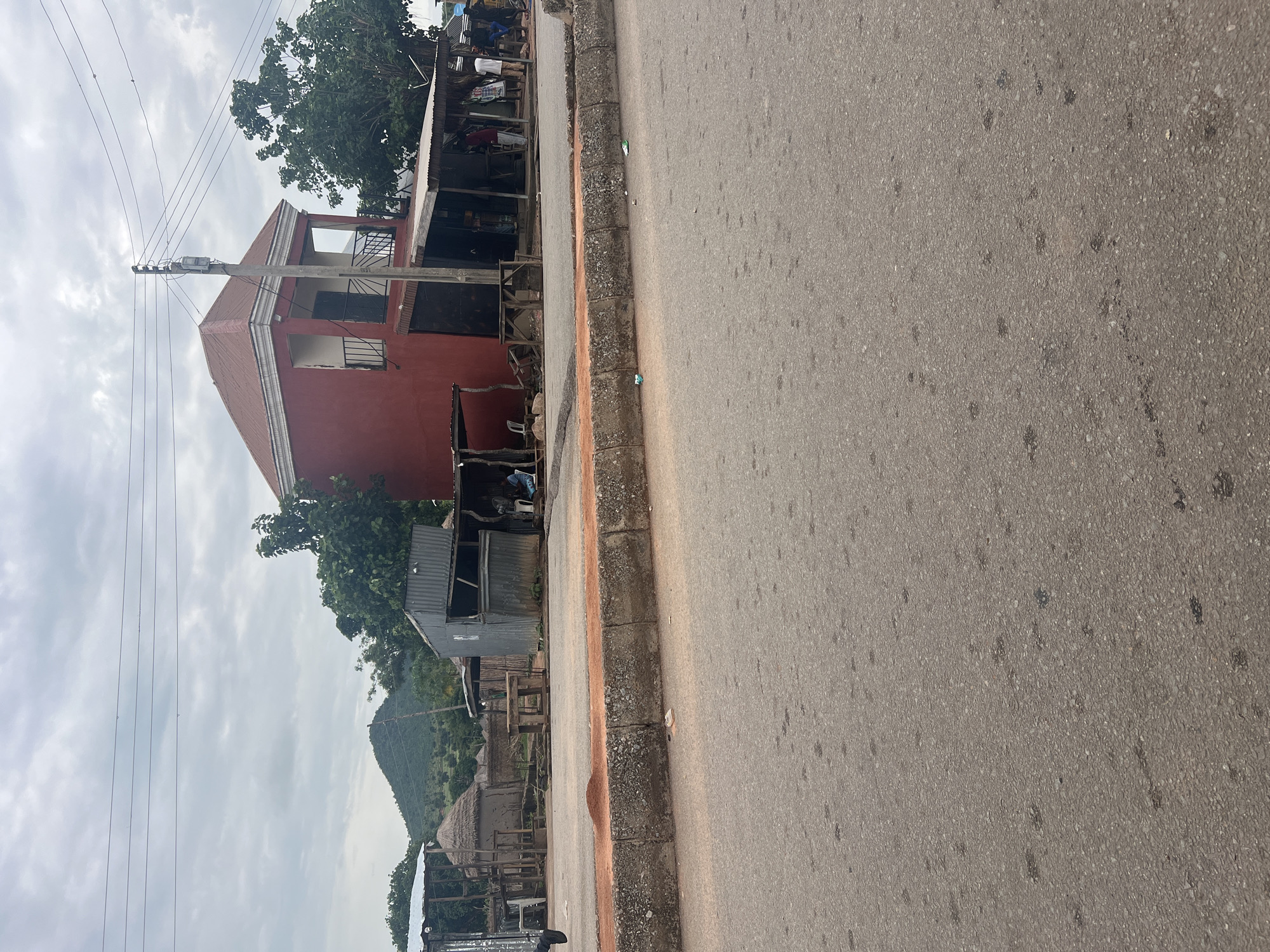 The image shows a street scene in Kona Garu, Nigeria. The scene is dominated by a two-story red building with balconies and windows. In front of the building are some smaller structures, possibly shops or shelters, made of corrugated metal and wood. There are a few people visible in the background, some tending to market stalls. The street itself is paved and has a distinct curb with rough texture. The sky is overcast, suggesting a cloudy day. In the distance, a lush green hill or mountain range is visible, adding a natural element to the otherwise urban landscape. Power lines run across the scene, indicating the presence of electricity infrastructure. The overall impression is of a busy, everyday scene in a Nigerian town, possibly during market hours.