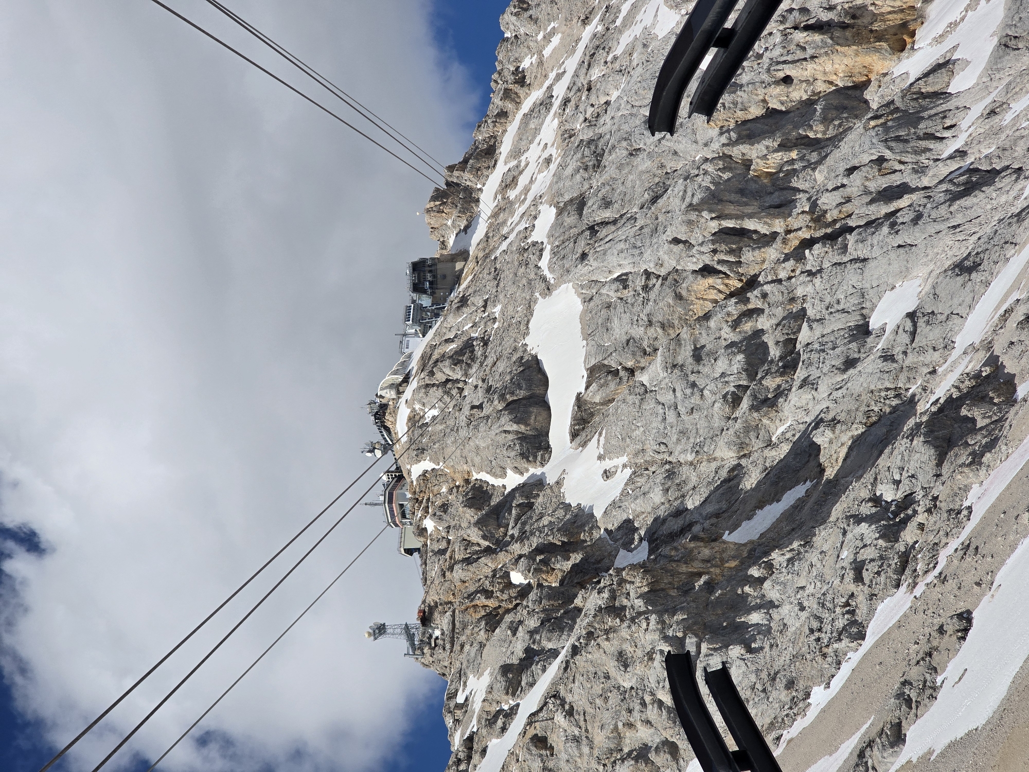The image shows a view of the Zugspitze mountain in Garmisch-Partenkirchen, Germany, from a cable car. The mountain is rocky with patches of snow. Several buildings, including a cable car station and a weather station with antennas, are visible on the summit. The sky is cloudy with patches of blue. The perspective suggests the viewer is in a cable car, looking up towards the mountain.