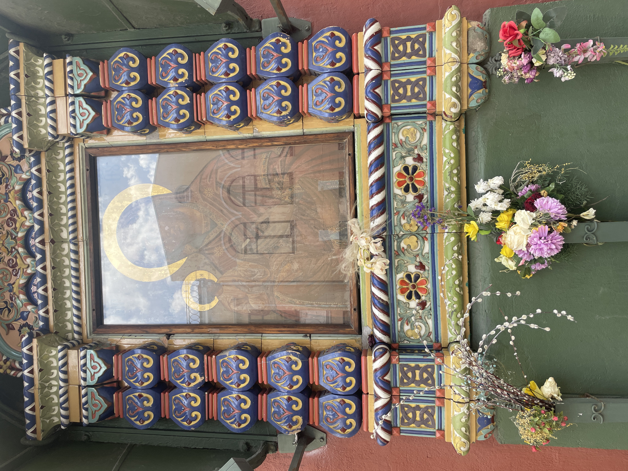 A close-up shows the exterior of a religious building, possibly a church, in Moscow, Russia, displaying intricate, colorful decorations and icons. A framed image sits in the center, reflecting the sky and a stylized golden crescent moon. The frame is surrounded by ornately carved and painted elements in blue, yellow, red, and green, including geometric patterns, floral motifs, and twisted columns. Fresh flowers and pussy willow branches are arranged around the architectural details. The exterior wall is painted green above and brick red below. Metal support brackets are also visible.