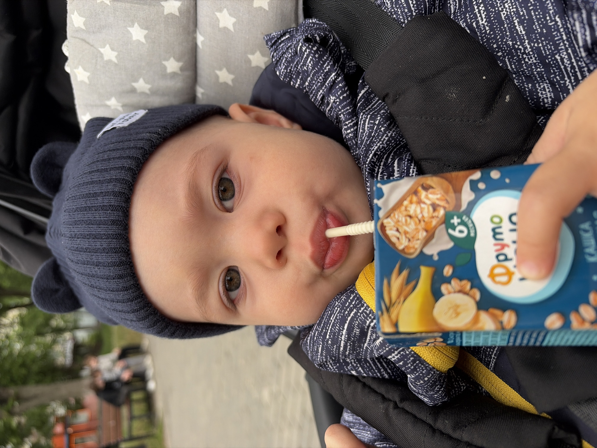 A baby wearing a dark blue knit hat with ears is drinking from a "Fruto Nyanya" juice box. The juice box features images of bananas and oats and indicates it is for babies aged 6 months and older. The baby is in a stroller with a grey and white star patterned liner. The person holding the juice box is partially visible. In the background, there is a park setting with people sitting on a bench. The image appears to be taken in the city of Chaikovsky, Russia, based on the branding on the juice box.