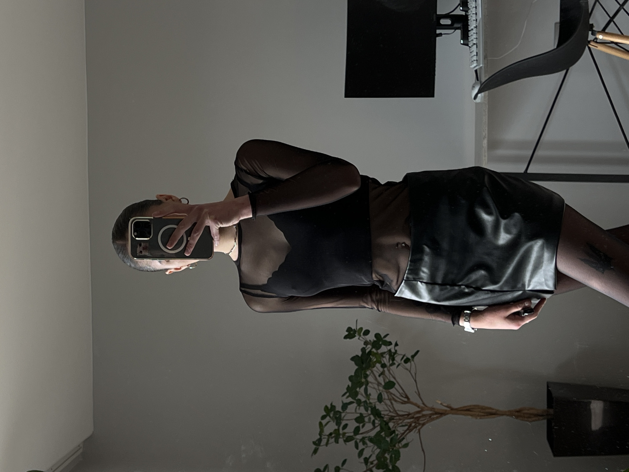 A person is standing in front of a mirror, taking a selfie. They are wearing a black sheer long-sleeved top, a black bralette, a black leather mini-skirt, and black patterned tights. The person has a belly button piercing. They are also wearing a silver watch and a ring on their left hand. To the left of the person is a tall potted plant. In the background, there is a desk with a computer monitor, keyboard, and mouse, and a black chair. The scene appears to be indoors, possibly a bedroom or dressing area. There is no visual information in the image to suggest the location is Ternopil, Ukraine.