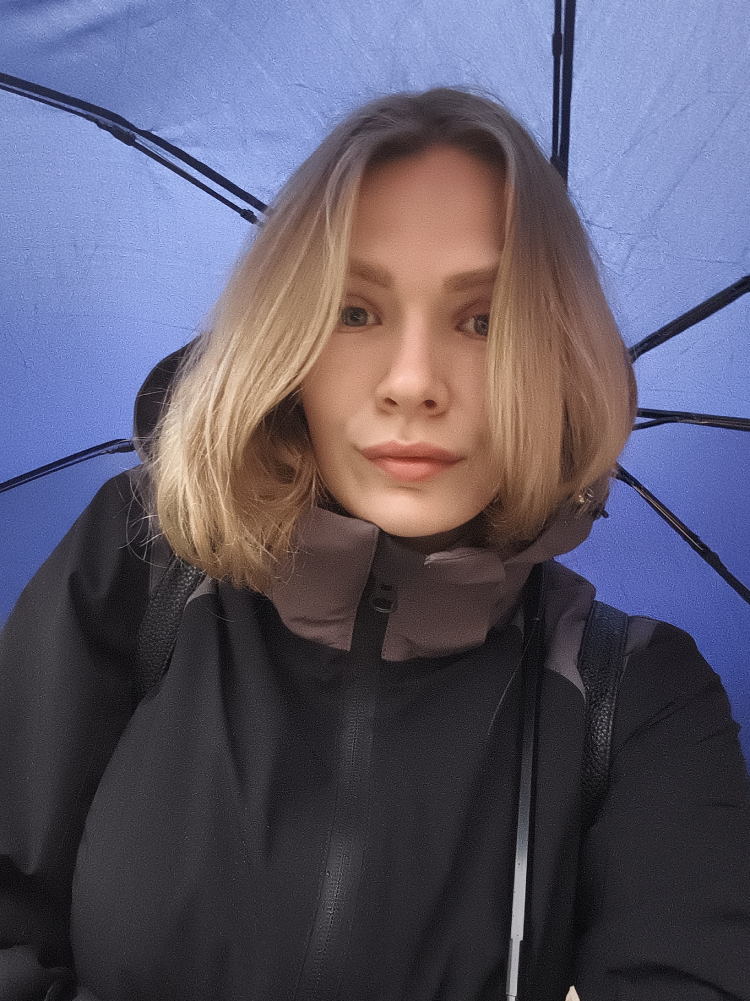 A woman is taking a selfie under a blue umbrella. She is wearing a black jacket with a gray collar and a black backpack. She has short blonde hair and light eyes. The location is not discernible as Izhevsk, Russia.
