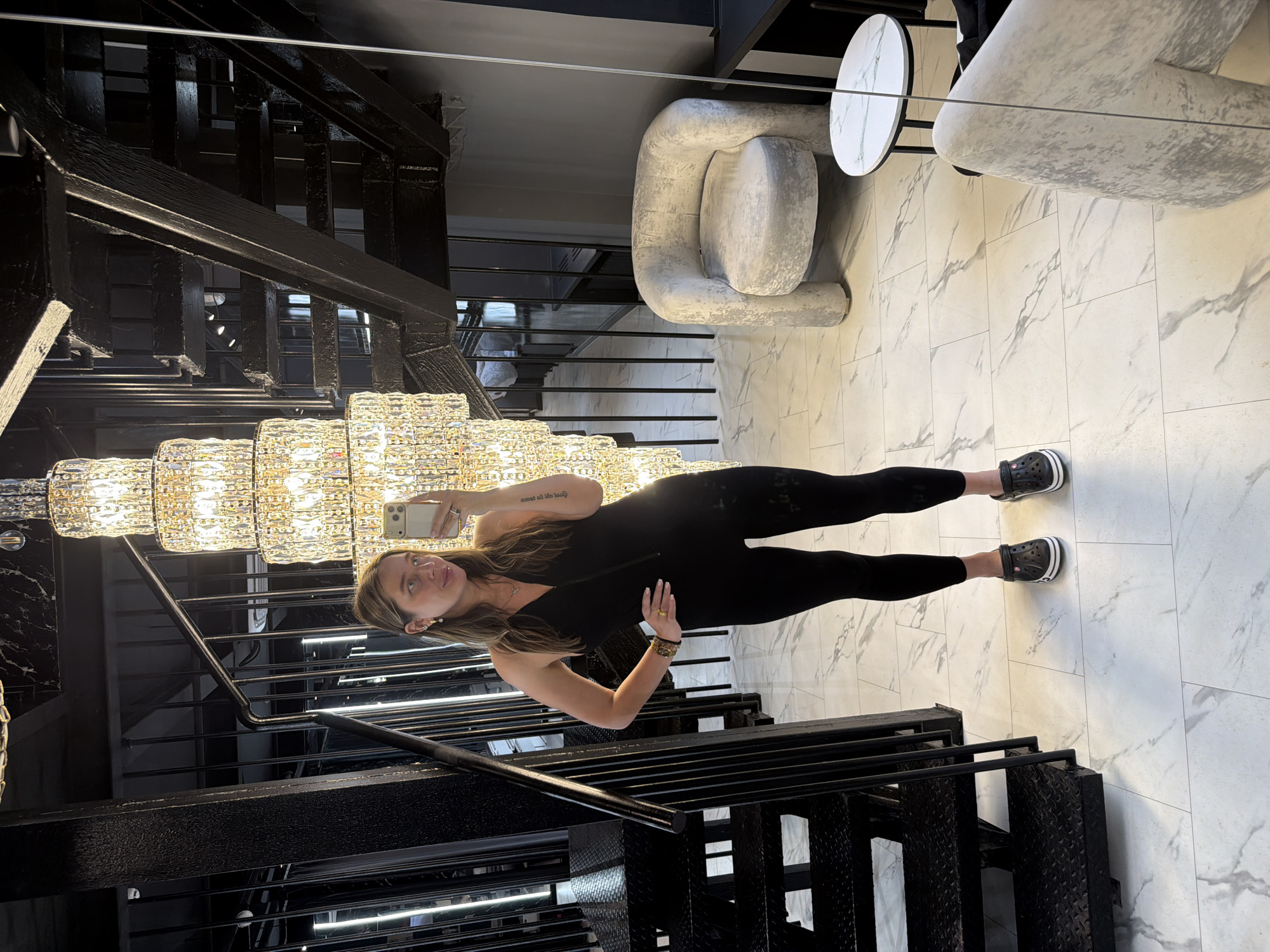 A woman is photographed in what appears to be an upscale interior location. She is holding a smartphone and taking a selfie, positioned near a staircase with black metal railings. A large crystal chandelier is visible above and behind her. The flooring is tiled in a light marble pattern. Furniture includes what looks like a modern chair and a side table. The location may be in Moscow, Russia, but this is uncertain. The overall impression suggests a fashionable or luxurious setting.