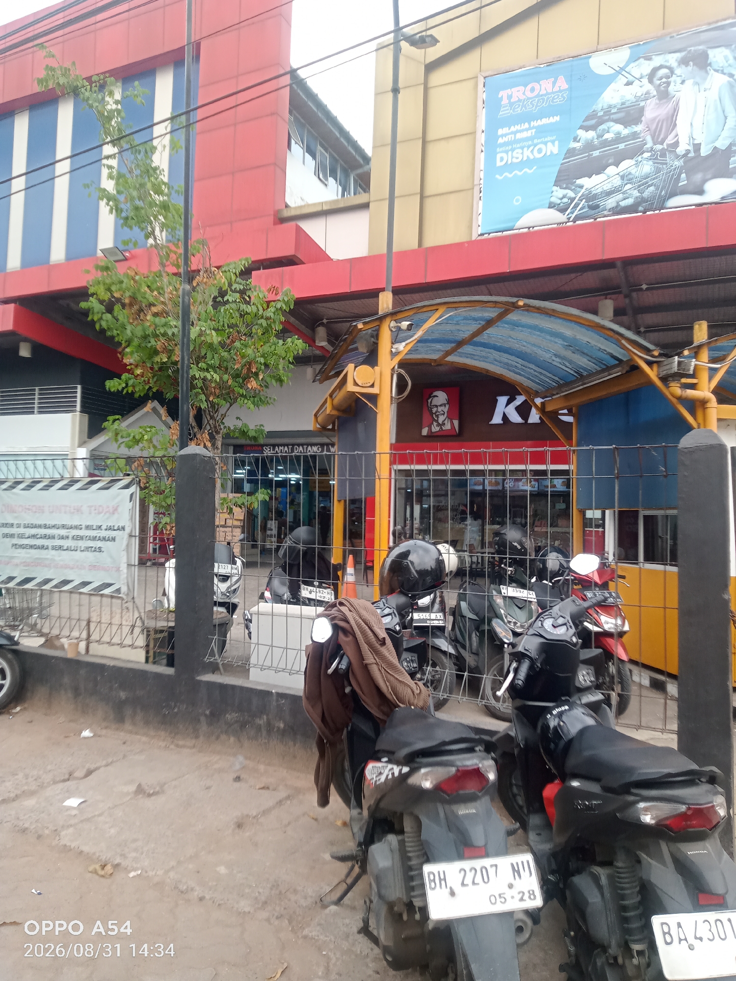 The image shows a KFC restaurant with a blue and yellow canopy in Jambi, Indonesia. Several motorcycles are parked in front of the restaurant, some with helmets and clothing on them. A red building with white and blue stripes is visible behind the KFC. A large billboard for "TRONA Ekspres" with an image of people shopping is displayed on the red building. A sign in Indonesian that reads "DILARANG UNTUK TIDAK PARKIR DI BADAN/BAHU JALAN MILIK JALAN DEMI KELANCARAN DAN KENYAMANAN PENGENDARA BERLALU LINTAS" is also visible, which translates to "DO NOT PARK ON THE ROAD/SHOULDER OF THE ROAD FOR THE SMOOTHNESS AND COMFORT OF TRAFFIC." The ground is paved and there is some debris on it. The timestamp "OPPO A54 2026/08/31 14:34" is overlaid on the image.