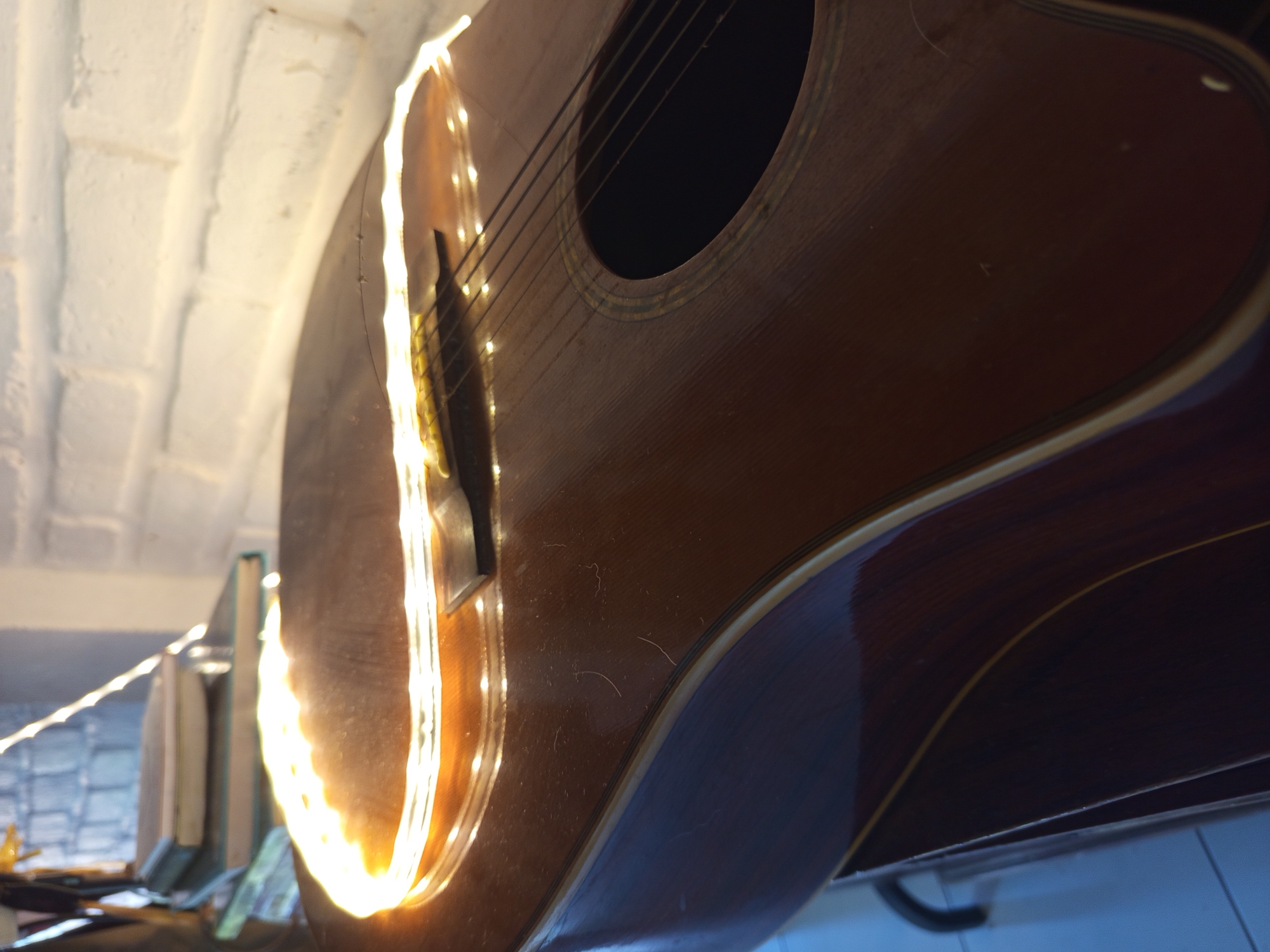 An acoustic guitar is prominently featured, positioned vertically or at a steep angle, adorned with a string of warm fairy lights that trace its left outline and extend into the background. The guitar itself is made of dark brown wood with a glossy finish, showing visible wood grain, fine dust, and minor surface imperfections. Its six strings are visible, stretching across the body towards the sound hole, which is encircled by a decorative rosette.
The bright, warm glow of the fairy lights illuminates the guitar and casts light onto the immediate surroundings. To the upper left, a white brick wall with an arched architectural detail is visible, brightly lit by the fairy lights. In the background, partially obscured, is a shelf or table holding stacked items, possibly books, and another section of wall with a lighter, possibly tiled pattern. The overall lighting suggests an indoor setting, likely during the evening or night, creating a cozy and ambient mood. There are no people visible, and no discernible text. The scene focuses entirely on the static display of the musical instrument.