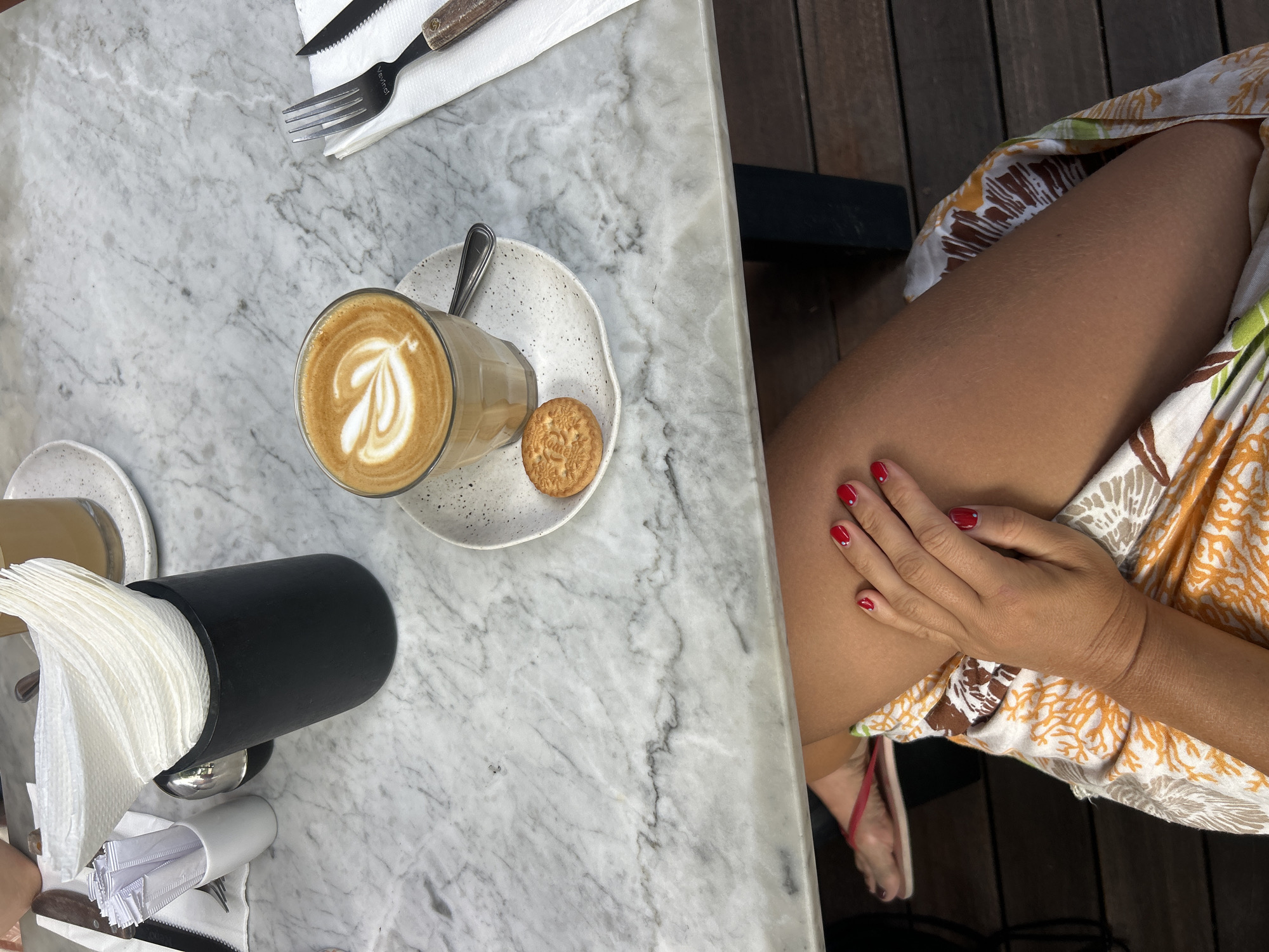 A close-up overhead shot captures a marble-topped table with various items, including a glass of latte art coffee, a small biscuit, a napkin holder, and cutlery. In the foreground, a woman's legs are visible, clad in a patterned skirt and red flip-flops. Her hand, with red-painted nails, rests on her skirt. The scene appears to be in Kuta, Indonesia, possibly at a cafe or restaurant.