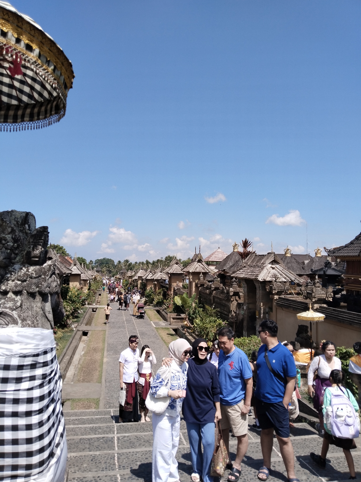 The image captures a scene at the Kehen Temple complex in Bangli, Indonesia, on a clear day. In the foreground, a large, intricately decorated umbrella with black and white checkered fabric and gold accents is partially visible on the left, casting a shadow. A stone statue, likely a guardian figure, is also present in the foreground, to the left of the umbrella.
The main subject is a pathway leading uphill, lined with traditional Balinese architecture featuring stone carvings and thatched roofs, some adorned with golden crowns. Numerous people are walking up and down the path. In the mid-ground, a group of tourists is gathered. Among them, a couple dressed in traditional Balinese attire, a man with a white shirt and a red sarong, and a woman with a similar sarong and a white top, stand out. Other individuals are in casual tourist clothing. Further up the path, more people are visible, some appearing to be locals in traditional dress and others tourists.
The environment is sunny with a bright blue sky and scattered white clouds. Lush green vegetation and trees are visible throughout the complex, interspersed with the stone structures. The pathway is paved with stones. The scene conveys a sense of cultural heritage and tourism activity within a temple setting.