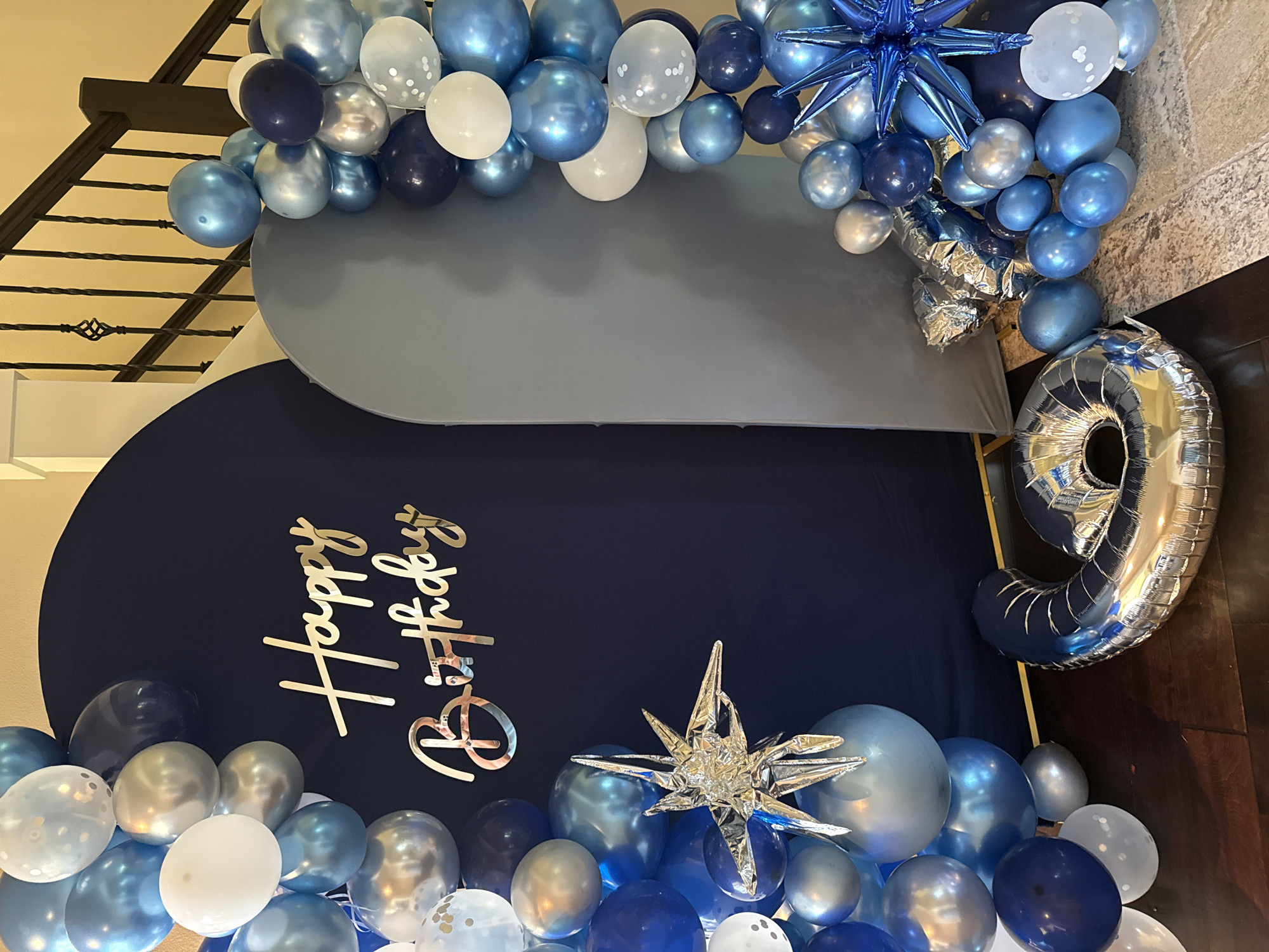 This image shows a birthday party setup in what appears to be a residential interior. The primary focus is a balloon arch and backdrop for a celebration. The backdrop is a dark navy blue fabric, possibly a stretched-out arch shape, with the words "Happy Birthday" written in a cursive, gold mirrored font. On the left side of the backdrop, there is a large cluster of balloons in various shades of blue, white, and silver. Some of these balloons are plain, while others have a subtle confetti or glitter pattern. A large, silver starburst decoration is also attached to the balloons on the left.
To the right of the navy backdrop, there is a large, light gray arch-shaped backdrop. More balloons, similar in color and style to those on the left, are arranged along the edge of this gray backdrop and spilling onto the floor. Scattered among the balloons on the floor are several foil balloons. One prominent foil balloon is shaped like the number "6" and is silver. Another is a large, silver ring or donut shape.
The floor is a dark wood in the foreground, transitioning to a patterned rug in the background. A staircase with a black wrought-iron railing is visible in the upper right corner of the image. The lighting suggests an indoor setting, likely illuminated by ambient room light. There are no people visible in the image. The setting does not provide specific indicators to place it in Vancouver, United States of America.