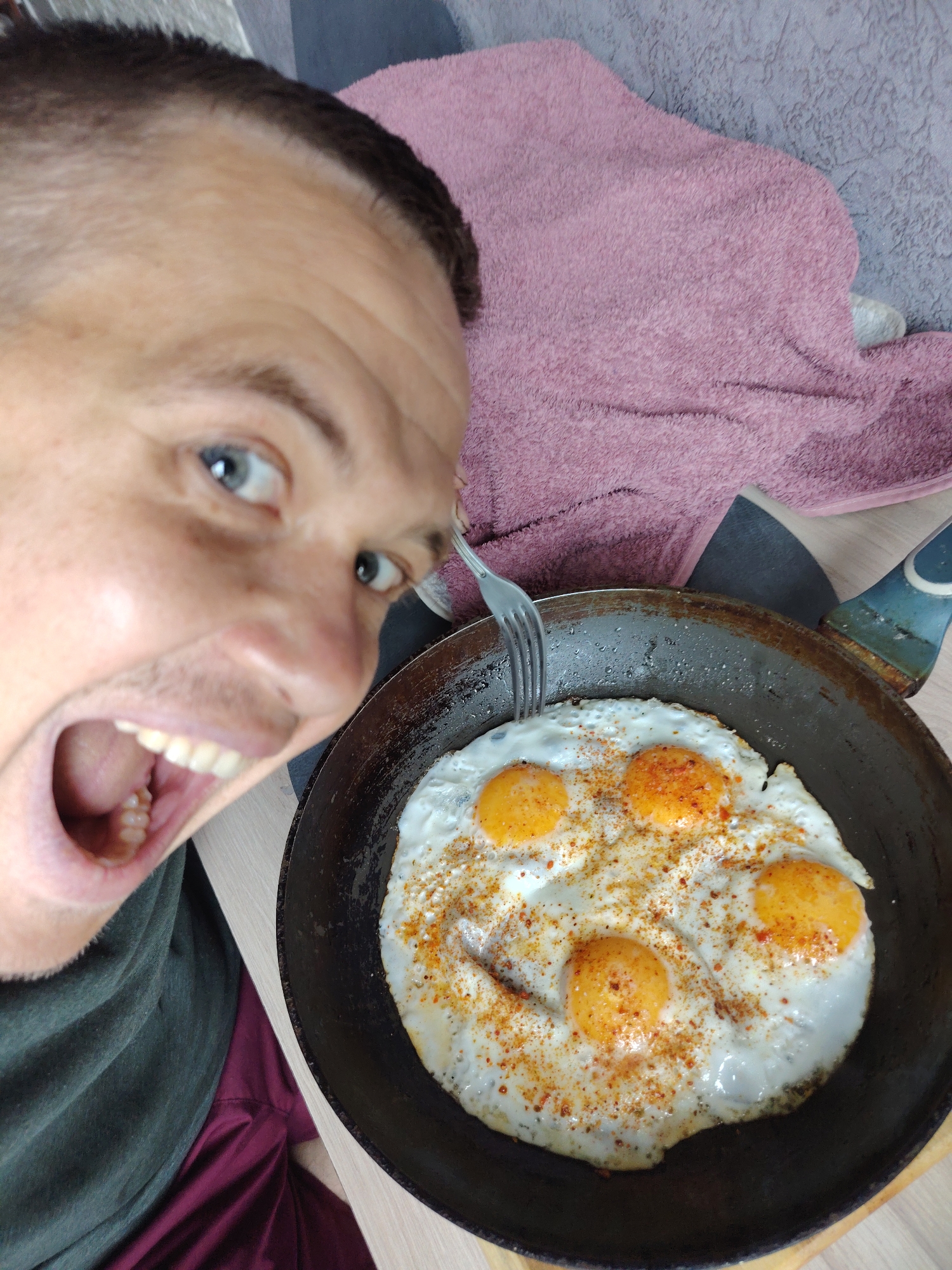 A man is looking into the camera with his mouth open. He has blue eyes and short brown hair. He is wearing a dark green t-shirt and maroon shorts. In front of him is a frying pan with fried eggs. The eggs are sprinkled with red pepper flakes. A pink towel is behind the man and the frying pan. The scene appears to be indoors, possibly in a kitchen.