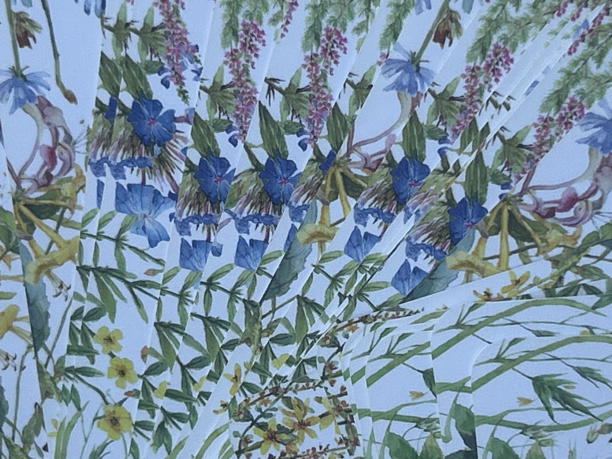 The image displays a close-up, downward-angled view of a white, pleated textile or paper surface featuring a repeating botanical pattern. The pattern is composed of various types of watercolor-style flora, including clusters of small yellow flowers, prominent blue funnel-shaped flowers, delicate light-blue multi-petaled flowers, sprigs of small pinkish-purple blossoms, and diverse green foliage with stems. The botanical elements are scattered densely across the light background, creating an all-over floral motif. The pleats run diagonally across the frame, approximately from the upper left to the lower right, creating distinct bands of light and shadow and segmenting the pattern. No people, animate subjects, or specific actions are depicted. The environment or specific location, including a city, cannot be determined from the image content.