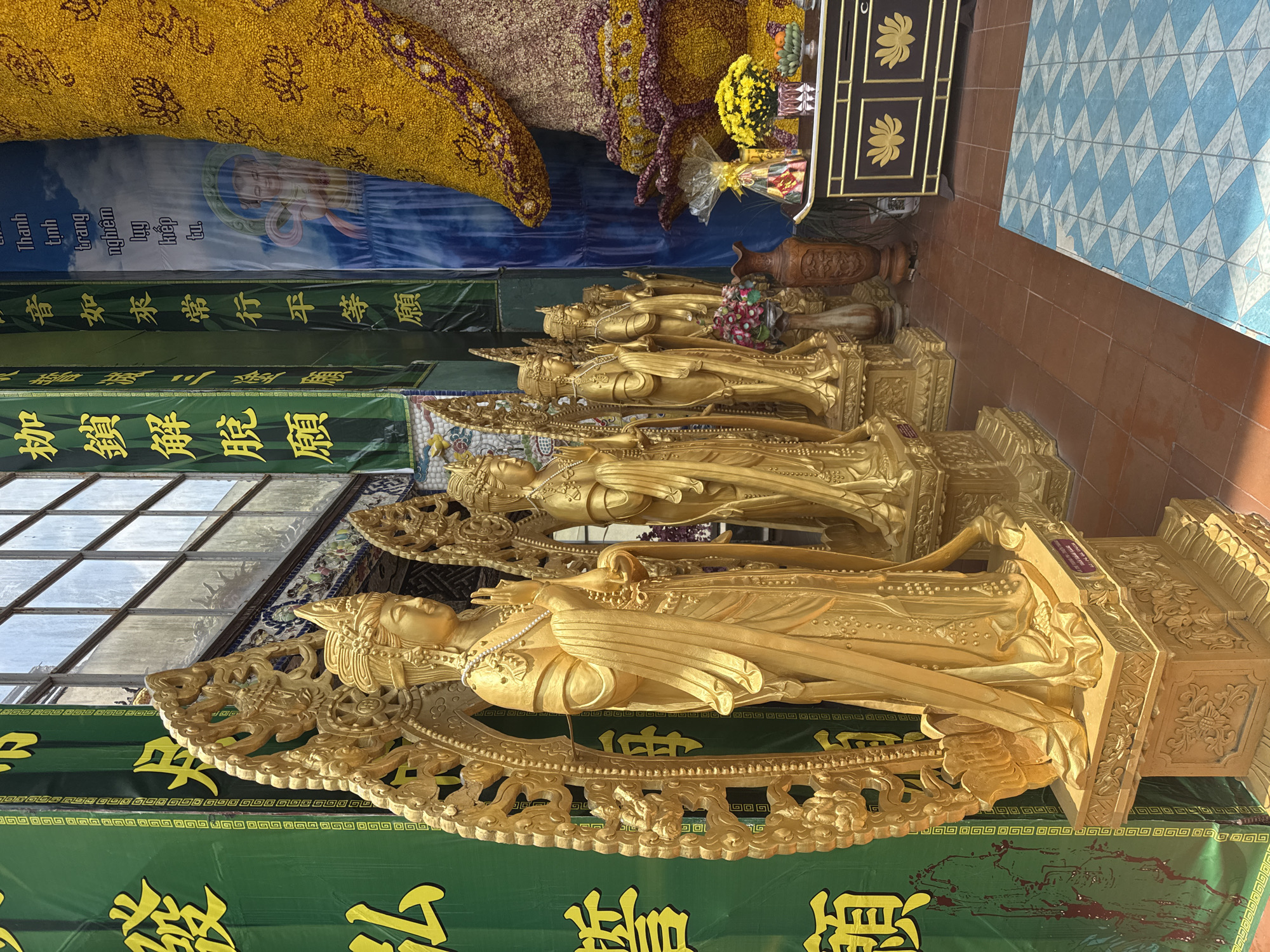 The image captures a scene within a temple, potentially located in Xuân Thành, Vietnam, though definitive confirmation from the image is not possible. A series of golden Buddha statues are prominently displayed, each intricately detailed and positioned in a row. Green banners adorned with yellow traditional script hang below the statues. Above the statues, a floral arrangement made of yellow flowers is visible, along with banners bearing text in both Vietnamese and Chinese. The temple floor is covered with a mix of tile patterns, and a brown cabinet stands nearby. The overall impression is one of reverence and ornate religious display.