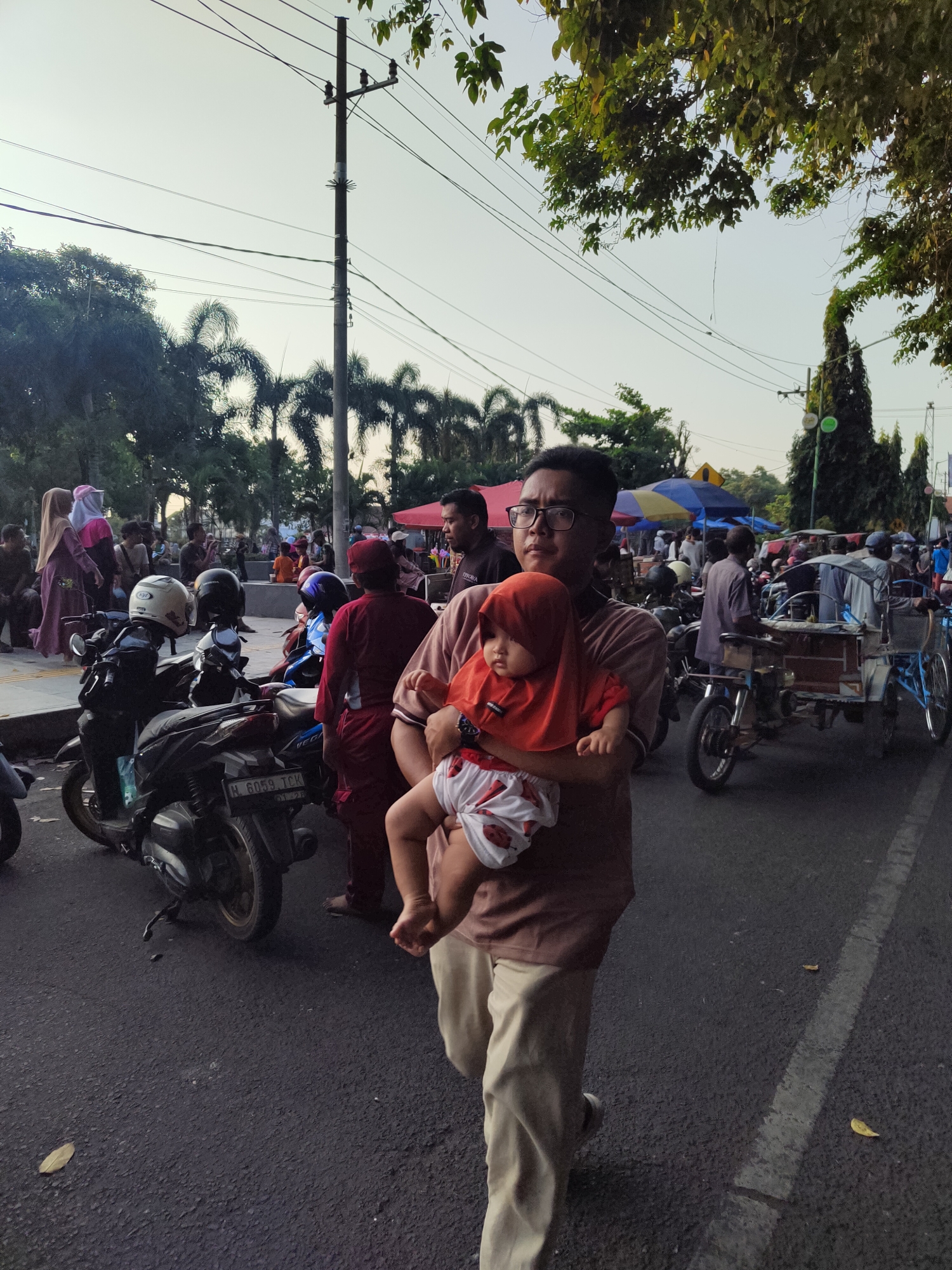The image depicts a street scene in Bangil, Indonesia, during daylight hours, likely late afternoon due to the warm lighting and long shadows. A man in the foreground is carrying a baby, who is wearing an orange headscarf and white and red patterned pants. The man is wearing a brown shirt and light-colored pants. In the background, a busy street is visible with various people, motorcycles, and a pedicab. There are trees and utility poles lining the street. The overall atmosphere appears to be that of a local market or gathering, common in Indonesian cities.