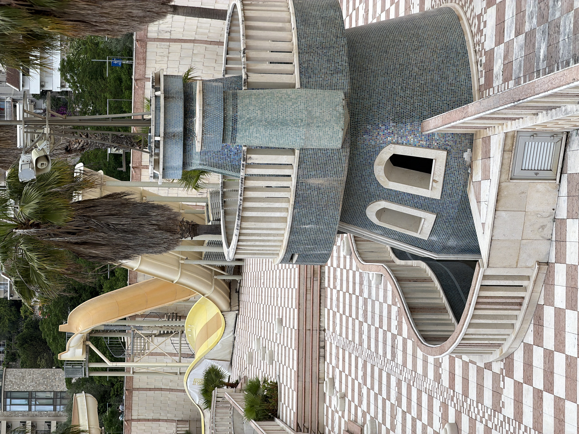 This media file captures an outdoor leisure or resort area, likely a water park or hotel complex, in Boreti, Montenegro.
The scene is dominated by intricate architecture and recreational facilities. Prominent on the left are light yellow/tan water slides, one with light brown and yellow stripes, winding downwards, supported by a metal framework. Adjacent to these are large palm trees with long, dry fronds, scattered across the area and in the foreground.
The central and right portions of the image showcase structures covered in small, square mosaic tiles, predominantly in shades of blue and green, suggesting water features or decorative elements. Integrated into these tiled structures are multiple sets of curving, white or light-colored stairs with graceful balustrades. Some structures feature arched window-like openings.
The ground and some vertical surfaces are covered with a distinctive checkerboard pattern of light-colored and reddish-brown tiles, with variations including smaller checkerboard borders. In the background, lush green trees and shrubs are visible, along with parts of other buildings. A tall metal pole equipped with floodlights and horn speakers stands on the left.
No people are visible, lending a sense of tranquility to the scene. The bright, even lighting indicates it is daytime with clear weather. A blue street sign with a white arrow pointing to the left is discernible in the distant background. The overall impression is one of a well-maintained, architecturally stylized leisure environment.