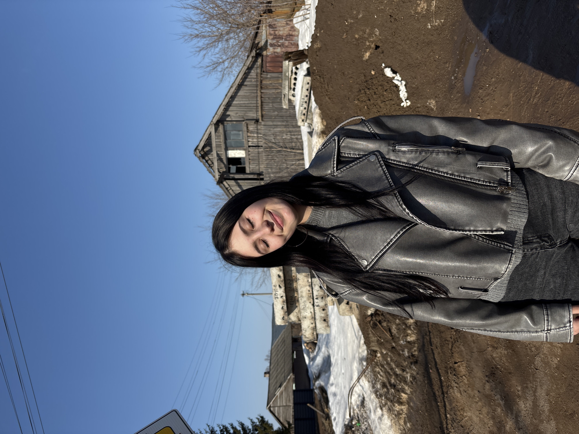 A young adult female subject with long dark hair is depicted outdoors in Фоки, Russia, under a clear blue sky. She wears a grey pseudo-leather jacket over a grey sweater and dark jeans. Her eyes are closed, and her head is slightly tilted, with a faint smile. Silver hoop earrings are visible. To her immediate left, a yellow diamond-shaped road sign is partially visible. The foreground consists of uneven, muddy ground with patches of snow and dirt mounds. Behind the subject, a stack of large, precast concrete slabs, some marked "7012-P" and "FPK 8612-P," is present. In the background, two weathered wooden structures are visible: a smaller house on the far left and a larger, two-story gabled barn or shed with an upper window opening and a lower metal door to the right. Bare deciduous trees are scattered in the background, along with a coniferous tree on the far left. Overhead power lines span the upper frame. The scene is brightly lit by direct sunlight, indicating an early spring environment.