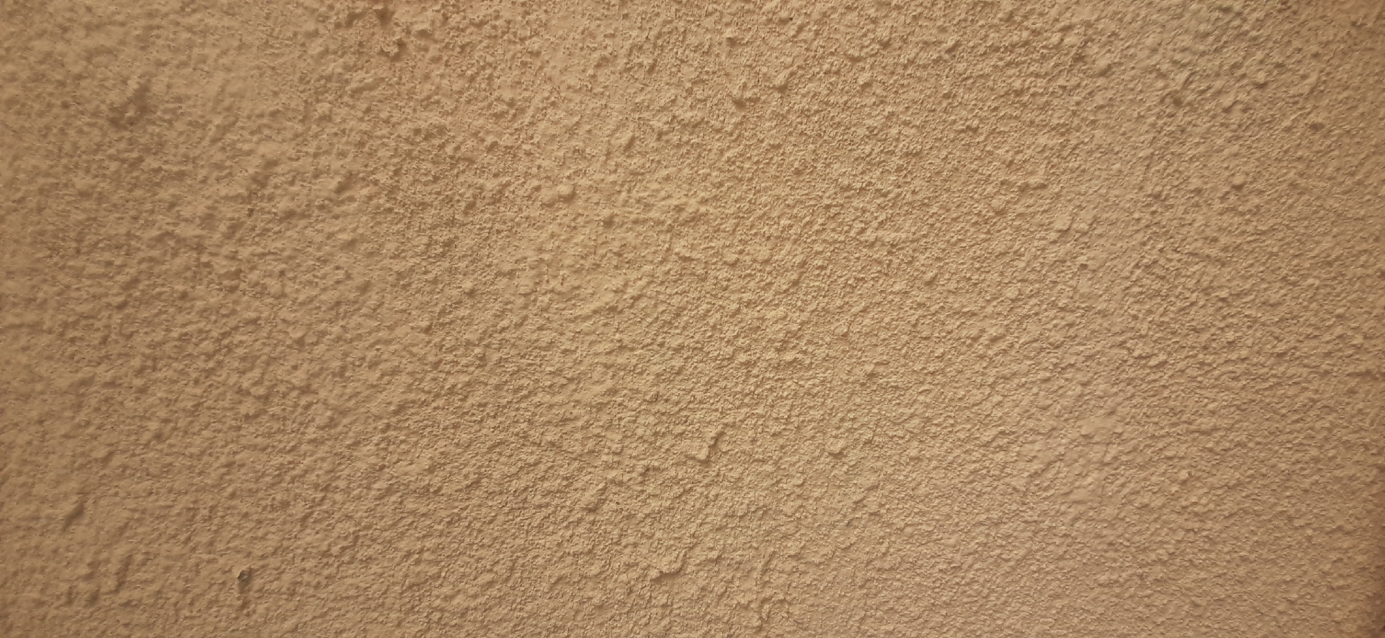 This is a close-up, full-frame, landscape-oriented photograph of a textured wall. The wall has a rough, stucco-like finish with a uniform, light brown or tan color. The texture is uneven, with many small bumps and pits across the surface, creating a subtle, dappled pattern of light and shadow. There are no people, objects, or clear indications of a specific scene or event. The lighting appears to be soft and diffused, suggesting it is not direct sunlight and could be indoors or an overcast day. There are no visible text elements in the image. The image focuses entirely on the texture and color of the wall, providing a neutral and abstract visual.
