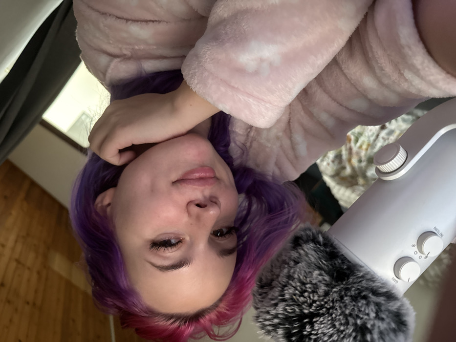 An upside-down, close-up shot of a woman with purple and pink hair, wearing a pink robe. She is positioned in front of a white microphone with a furry windscreen. The microphone's gain and pattern knobs are visible. A wood panel and window are in the background. The lighting is soft. The woman's expression is relaxed and slightly smiling.