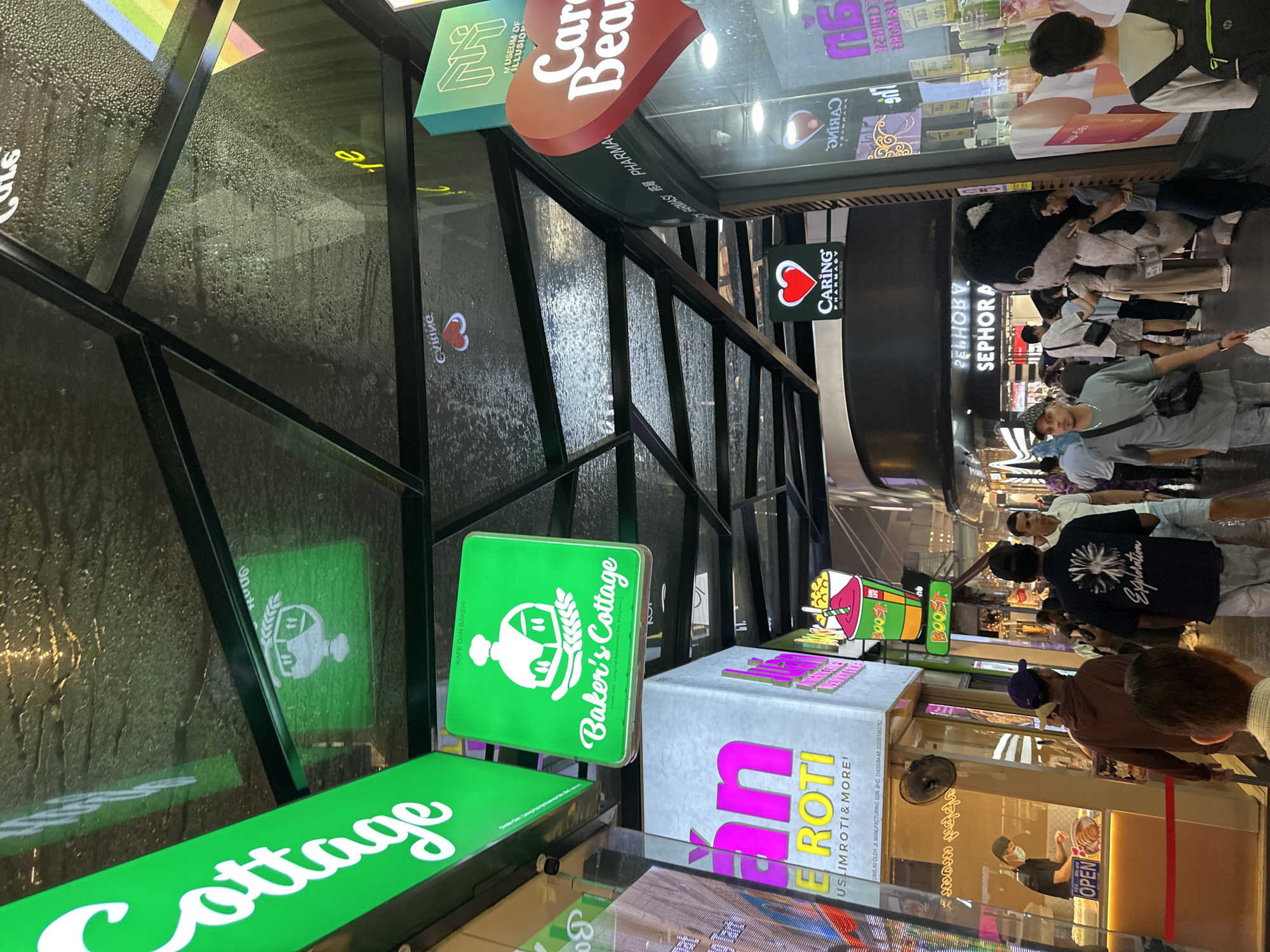 The image captures a bustling indoor shopping or commercial corridor, likely within a mall in Kuala Lumpur, Malaysia. The perspective is slightly elevated, looking down into a passage filled with people and brightly lit shopfronts.
Several individuals are visible, walking, standing, and browsing the shops. In the foreground, a person in a light green shirt and a cap is seen. Further into the corridor, other shoppers include one holding a large black and white plush toy. Another person, wearing a black shirt, is observed behind a counter with an "OPEN" sign.
The scene is dominated by numerous illuminated business signs. Prominently on the left are green signs for "Baker's Cottage," featuring a logo of a baker's hat and wheat, accompanied by "KAFE DAN BAKERI" (Cafe and Bakery). Another sign beneath reads "Cottage." Next to this, a large light box displays "an EROTI" and "USLIMROTI & MORE!", along with manufacturing details including "DIMILIKI OLEH JL MANUFACTURING SDN. BHD. (1400084-M 202001043762".
Further down the corridor, multiple green "BOOST" juice bar signs are visible. Closer to the right, a "CARING PHARMACY" sign with a distinct red heart logo is prominent. Reflections on the glass facades show reversed text, including "SEPHORA" and "Y PURUASI 药局 PHARMA." Other partially visible signs include "MUSEUM OF ILLUSION" and "Car Bear" (likely "Care Bear"). Some glass panels along the corridor show visible condensation or water droplets. The time of day appears to be evening or nighttime, indicated by the artificial lighting. The overall atmosphere suggests a lively and active retail environment.