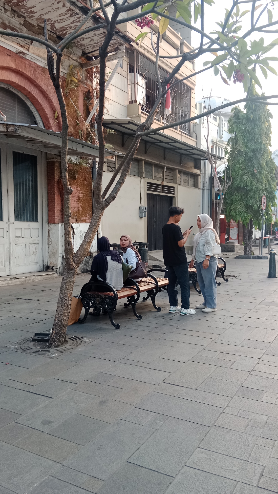 The image depicts a street scene in the historic Kota Lama area of Semarang, Indonesia. In the foreground, a man in a black t-shirt and dark jeans is standing and looking at his phone, while a woman in a white hijab and light blue jeans stands facing him, appearing to be in conversation. To their left, two women wearing hijabs are seated on a brown wooden bench with ornate black metal legs. One woman on the bench is looking towards the standing couple, while the other is looking away. The bench is situated on a paved sidewalk made of grey stone tiles. A young tree with thin branches is positioned in front of the bench, partially obscuring the seated women. In the background, a building with a red brick facade and arched windows is visible, alongside other structures with white and beige walls. The overall atmosphere suggests a casual daytime encounter in an urban environment with historical architecture.