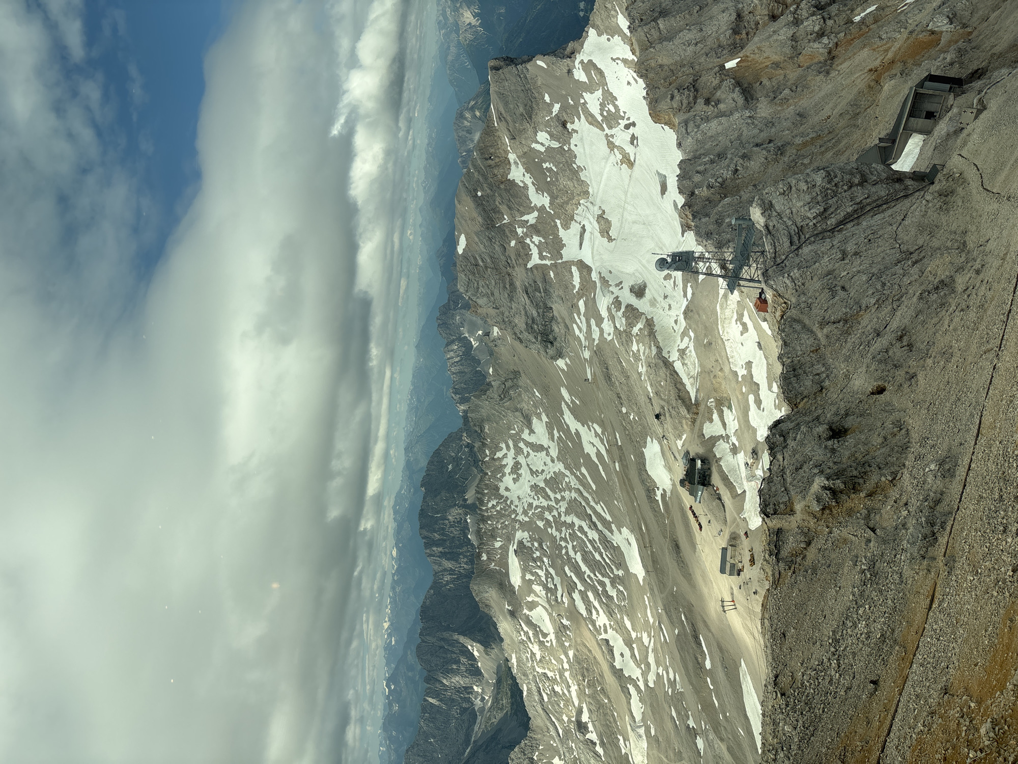This image is a panoramic view of a mountainous landscape, likely taken from a high vantage point. The foreground features a rocky, uneven terrain with sparse vegetation. In the mid-ground, a vast mountain range unfolds, characterized by steep, rugged peaks and valleys. Patches of snow are visible on the slopes, indicating a high altitude. Several man-made structures are present, including buildings, ski lifts, and a prominent radio tower with antennas, all appearing to be built into or on the mountainside. The background reveals a layered landscape of more distant mountains fading into a hazy horizon under a cloudy sky with patches of blue. The lighting suggests it is daytime. While the prompt mentions Ehrwald, Austria, the visual elements are consistent with a high alpine environment. There are no individuals or discernible actions or interactions depicted in the image.