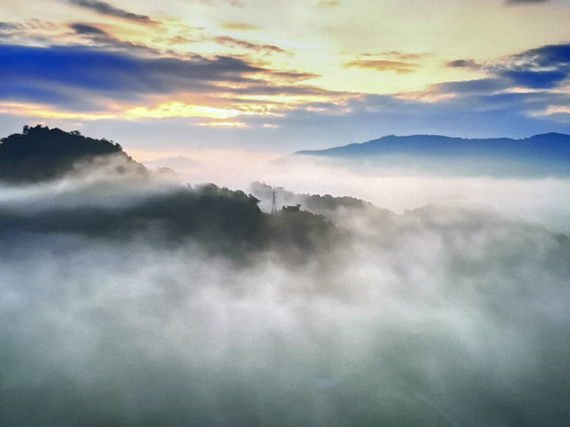 The image is a landscape photograph of mountains covered in fog, with a colorful sunrise in the background. The sky is filled with clouds, some of which are illuminated by the rising sun, creating hues of orange, yellow, and blue. The mountains are shrouded in a thick layer of mist, with the peaks of some emerging from the fog. The foreground is dominated by the dense fog, which obscures the lower parts of the mountains. There are no people or specific man-made structures visible in the scene. The image evokes a sense of tranquility and natural beauty.
There is no indication that this image depicts the city of Burgas, Bulgaria. The landscape features are consistent with mountainous regions where fog is common, particularly during sunrise or sunset.