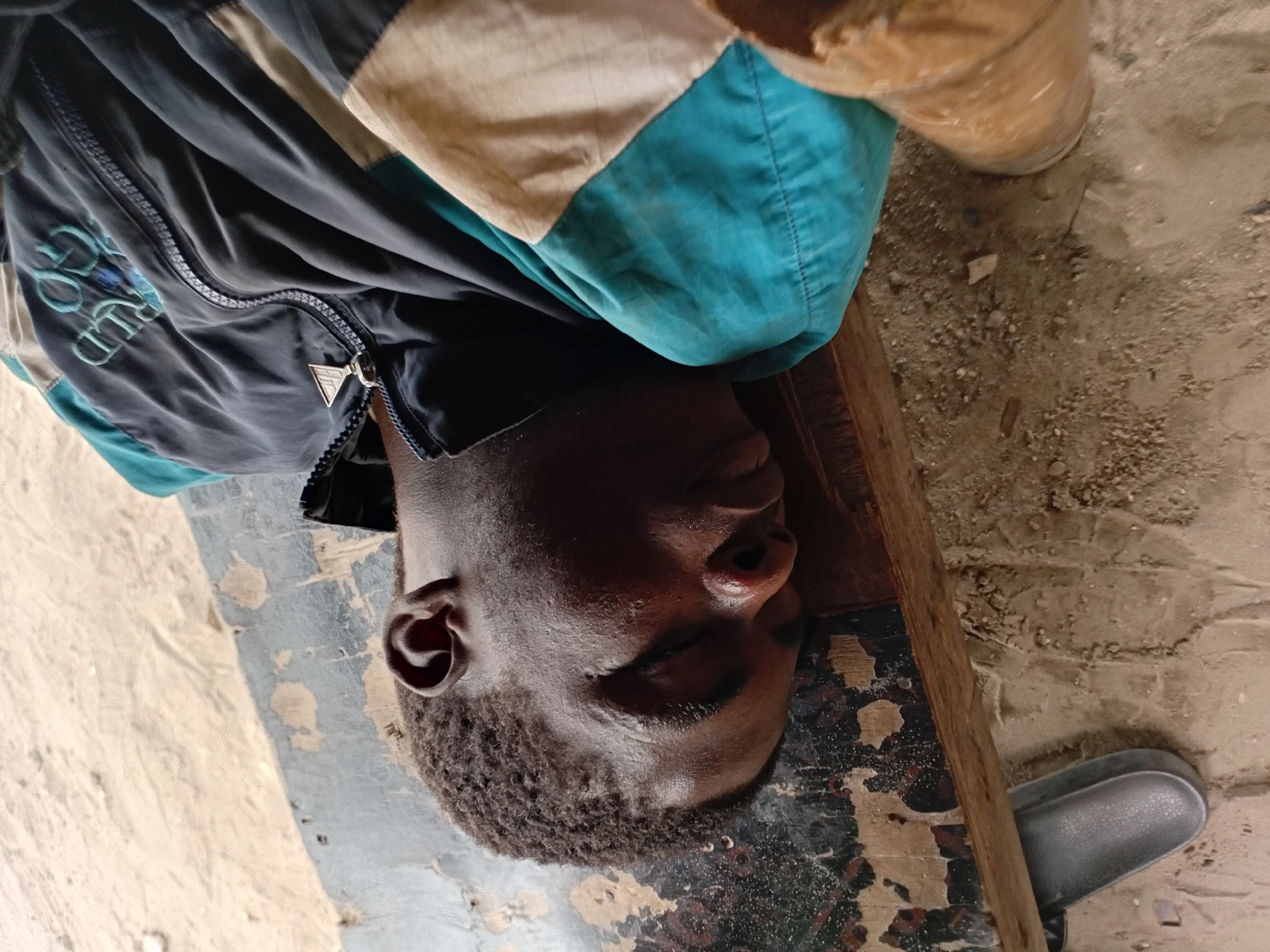 A young Black male is lying face down, asleep on a worn wooden bench. He is wearing a dark jacket with teal and tan accents, and a zipper is visible in the front. The bench is old and appears weathered, with chipped paint and exposed wood. The surrounding ground is sandy and dusty. A grey sandal is partially visible beneath the bench. The scene appears to be outdoors, possibly in a public space given the bench. The lighting suggests it is daytime.