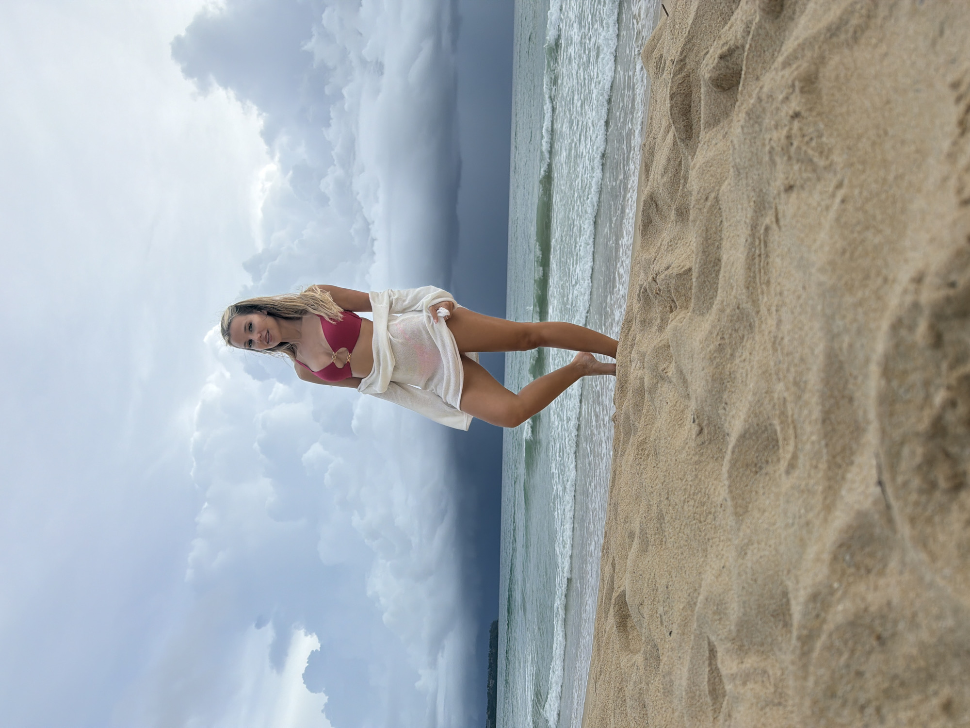 A young woman with blonde hair is pictured on a sandy beach. She is wearing a pink bikini and a white cover-up. The woman is leaning on her hands and looking at the camera with a smile. The background shows a stormy sky with dark clouds on the left and a bright sky with white clouds on the right. The ocean is visible to the left of the woman, with waves rolling onto the shore. The sand is light brown and textured, with footprints visible. The scene appears to be taking place during the day, possibly before or after a storm. The location is identified as Choeng Thale, Thailand.