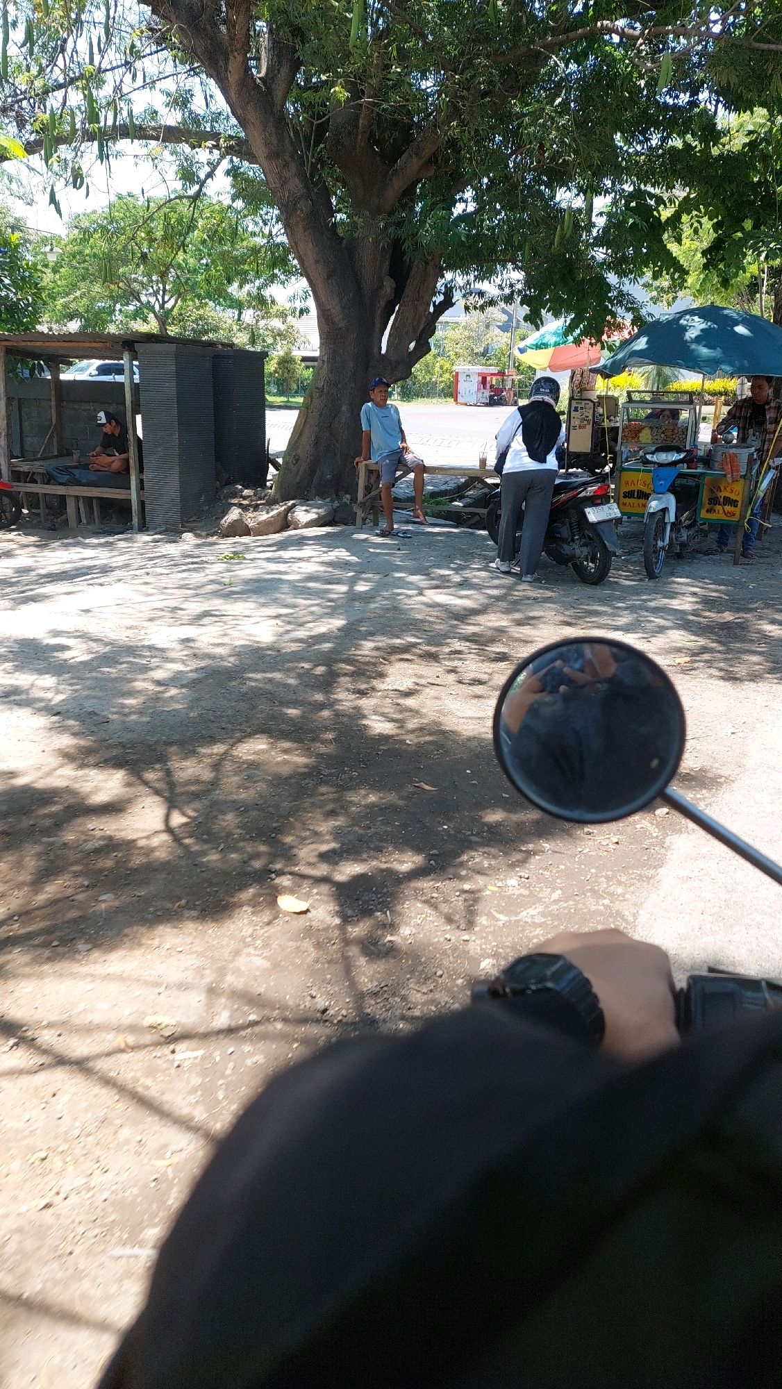 The image captures a scene in Siwalan Panji, Indonesia, under the dappled shade of a large tree. In the foreground, a person's hand with a black watch is visible, holding what appears to be a motorcycle handlebar, indicating the perspective of someone on a vehicle. To the left, a small, open-sided structure with a corrugated metal roof provides shelter. Inside this structure, a person is lying down, possibly resting. Near the large tree, a young man wearing a blue t-shirt and a blue cap is seated on a wooden bench. Further back, two people are standing near a food cart, one in a white shirt and black headscarf, the other partially visible in a checkered shirt. The food cart has umbrellas providing shade and displays various food items. Two motorcycles are parked near the cart. The ground is composed of dirt and pebbles, with shadows from the tree and leaves casting patterns. The environment suggests a roadside or a public gathering area.