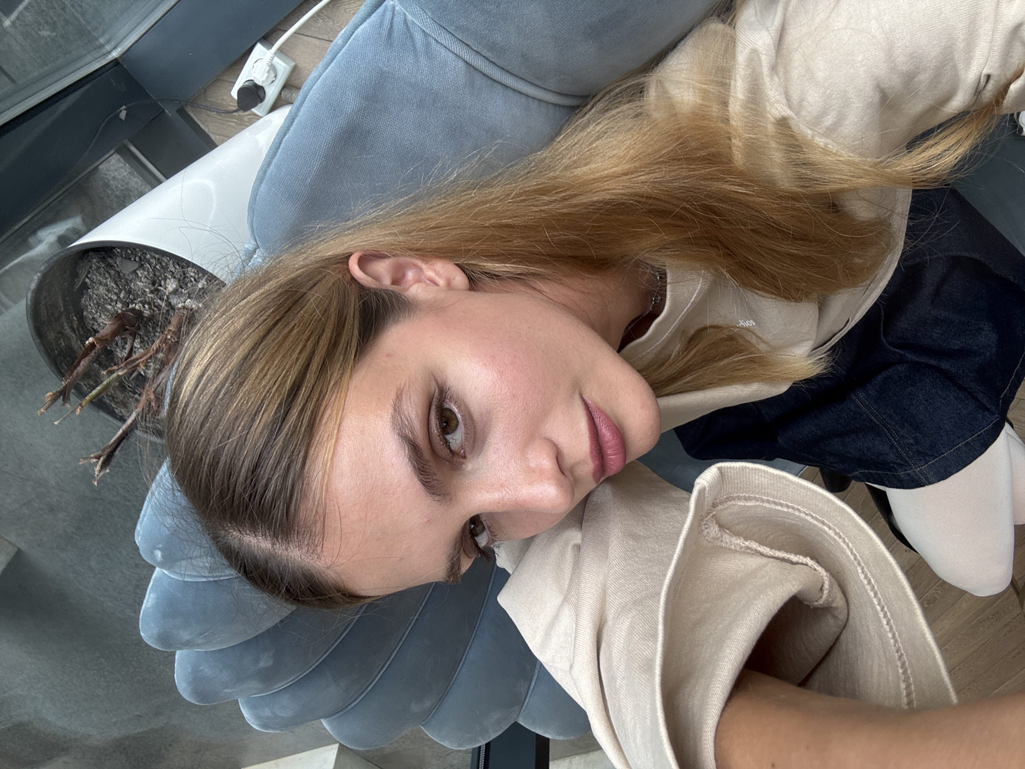 A young woman is lying on a blue scalloped chair, angled to the left, indoors, possibly in Tbilisi, Georgia, judging by the type of architecture and interior design common to the region. The subject has fair skin, light brown hair with blond highlights, and is wearing a light beige top with dark denim or black fabric and white tights or socks. The environment includes a potted plant in a white container and what appears to be a glass and dark colored wall or window behind the chair. There's an electrical outlet on the floor near the potted plant.