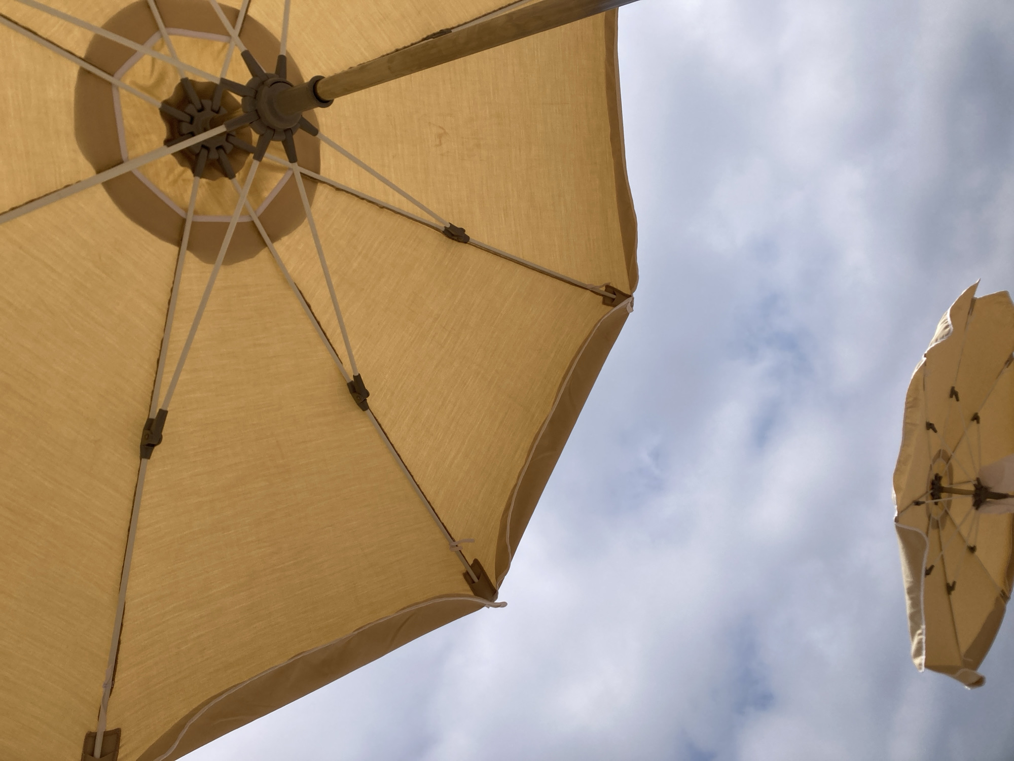 An upward shot of two yellow beach umbrellas against a cloudy sky. The main umbrella is in focus, taking up the majority of the frame. Its fabric has a linen-like texture and is supported by a metal frame with several spokes. The center of the umbrella has a darker, circular shadow cast by the mechanism. The second umbrella is smaller and out of focus in the lower portion of the image. The sky is filled with soft, grey and white clouds, suggesting an overcast day. The scene appears to be on a beach, common in coastal areas like Lloret de Mar, Spain. No people are visible.