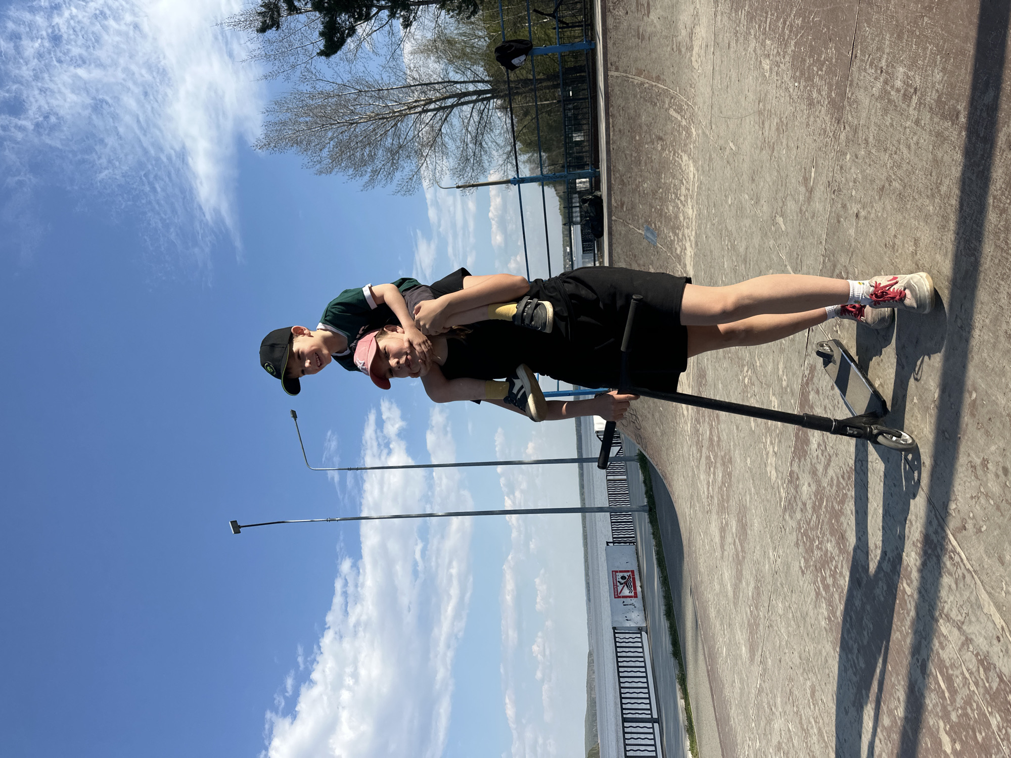 Two children are present in the image. The older child, wearing a black dress and a pink cap, is standing on a scooter on what appears to be a skate park surface. The younger child, sporting a green shirt and a black cap, is riding piggyback on the older child. The backdrop includes a body of water with a fence, street lights, trees, a blue sky with scattered clouds, and part of the structure of the skate park. Based on the location information provided, the scene is likely situated in Chaikovsky, Russia.