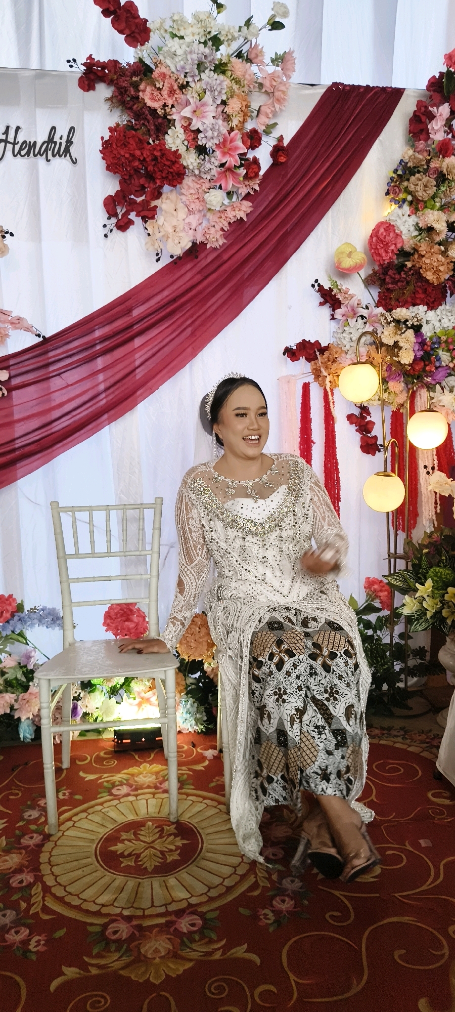 A full-body, vertical, outdoor shot shows a woman in a traditional white, beaded wedding gown and a patterned skirt seated on a white chair at a wedding reception in South Tangerang, Indonesia. She is smiling broadly and looking to her right. The bride's hair is pulled back in a bun and she is wearing a tiara. Behind her is a white backdrop decorated with a deep red draped fabric, floral arrangements in red, pink, and cream colors, and string lights. The floor is covered with a red carpet featuring a floral circular medallion in the center. The chair is a simple white cross-back design. To the bride's left, there are more floral decorations. The lighting is bright and appears to be natural, with additional artificial lighting from the string lights and a floor lamp to the right of the bride. The overall scene conveys a festive and celebratory atmosphere.
