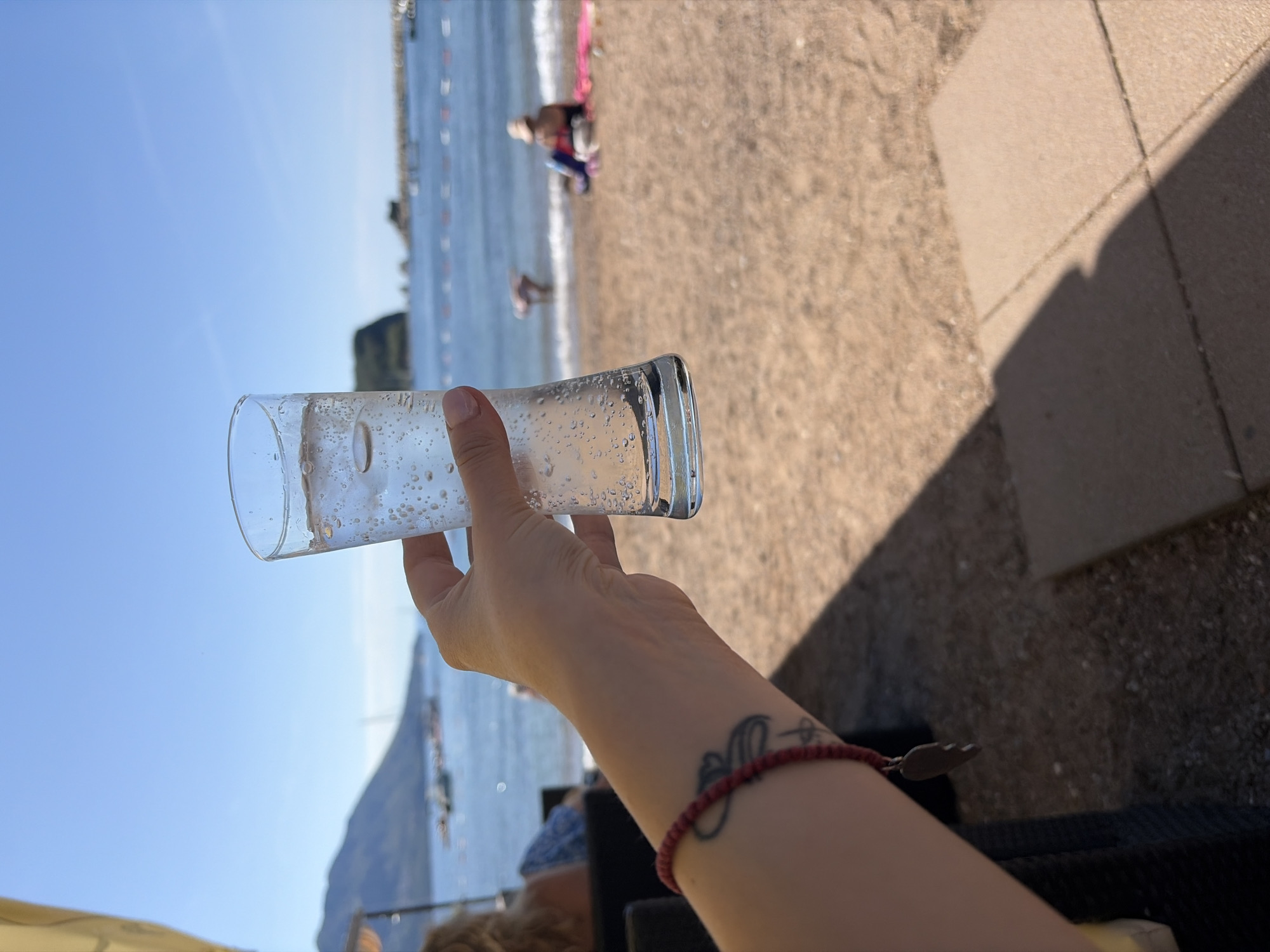 A first-person perspective captures a sunlit beach scene in Budva, Montenegro. In the foreground, a fair-skinned right arm holds a clear, ergonomically curved glass filled with a bubbly, clear liquid and ice cubes, positioned centrally. The arm wears a red beaded bracelet with a silver-colored, wing-shaped charm and displays a dark, stylized tattoo on the inner forearm.
The midground features a light brown sandy beach extending to the deep blue Adriatic Sea. Two individuals are visible on the beach: one person sits on a colorful towel on the sand to the right, wearing a hat, and another person stands in the shallow water near the gentle waves to the left. A line of red buoys marks a swimming area in the sea. Numerous small boats are anchored further out in the bay.
The background presents a clear, bright blue sky above a prominent green-covered mountain range on the left. To the right, the distinctive silhouette of Sveti Nikola Island, along with smaller rocky islets, rises from the sea. Shadows are cast on paved ground, likely concrete tiles, in the lower right, indicating an outdoor seating area. A sliver of a yellow canopy or parasol is visible in the upper left corner.