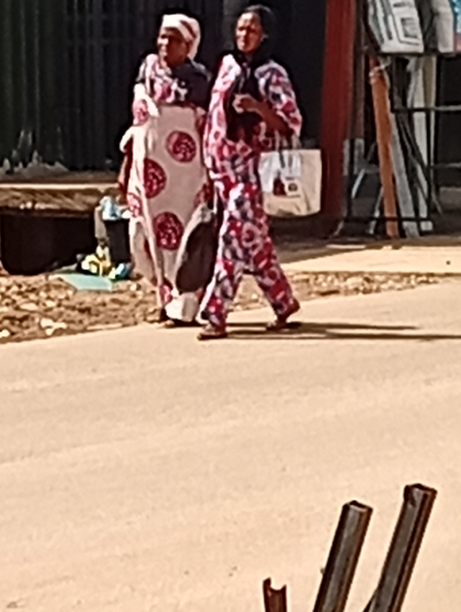 In Maiburiji, Nigeria, two women are walking side-by-side on a paved road. The woman on the left wears a patterned white and red dress and a white head covering. She carries a dark-colored bag on her shoulder. The woman on the right wears a similarly patterned red and white ensemble, including pants, and carries a white tote bag. They are walking away from a metal fence and a building facade. The road surface appears to be worn. In the foreground, dark-colored metal bars are partially visible. The lighting suggests it is daytime.