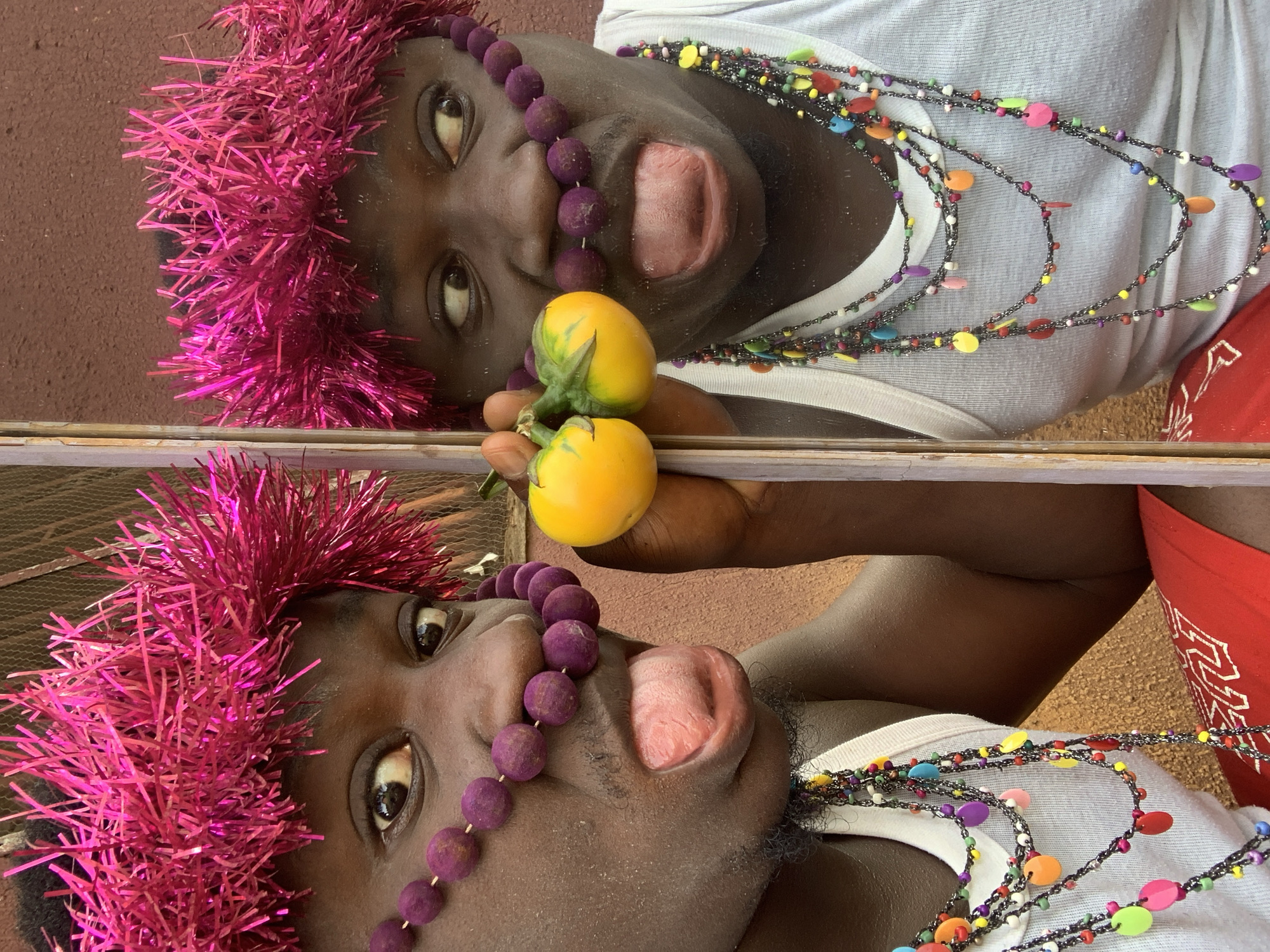 A man wearing a pink tinsel crown, purple bead necklaces around his nose and neck, and a white tank top poses with two yellow tomatoes against a brown textured wall. He is sticking his tongue out in the reflection of what appears to be a mirror or window. The scene is set in Akwanga, Nigeria. The man has a playful expression, and the vibrant colors of his accessories contrast with the muted background.