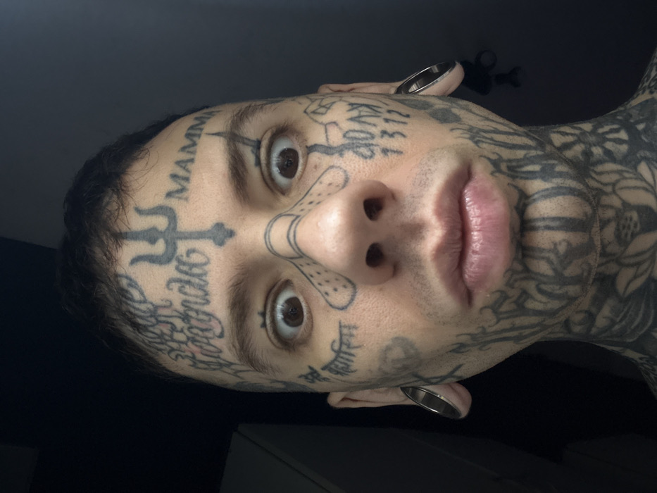 The image shows a close-up of a man's face covered in tattoos. The tattoos are primarily black ink and depict various symbols, words, and patterns. He has stretched earlobes with rings. His eyes are wide and his lips are full. The lighting is somewhat dim. Part of his shoulders and chest are also visible. The image is oriented with his face at an angle.