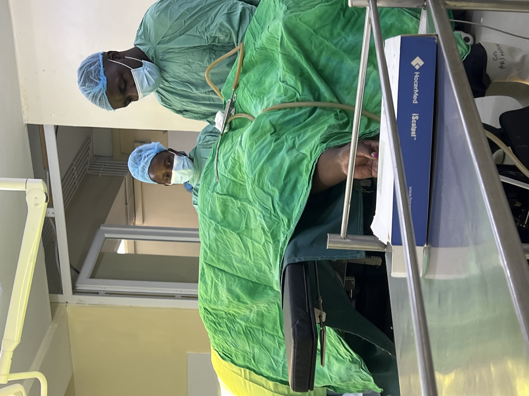 The image appears to show an operating room in Nairobi, Kenya, with two surgeons present. Both surgeons are wearing surgical scrubs, surgical caps, and face masks. The patient is draped with green surgical cloths. Surgical instruments and equipment, including tubing, are visible on and around the draped area. A box for a HocerMed iScalpel is on a metal cart near the patient. The environment is a brightly lit room with medical equipment.