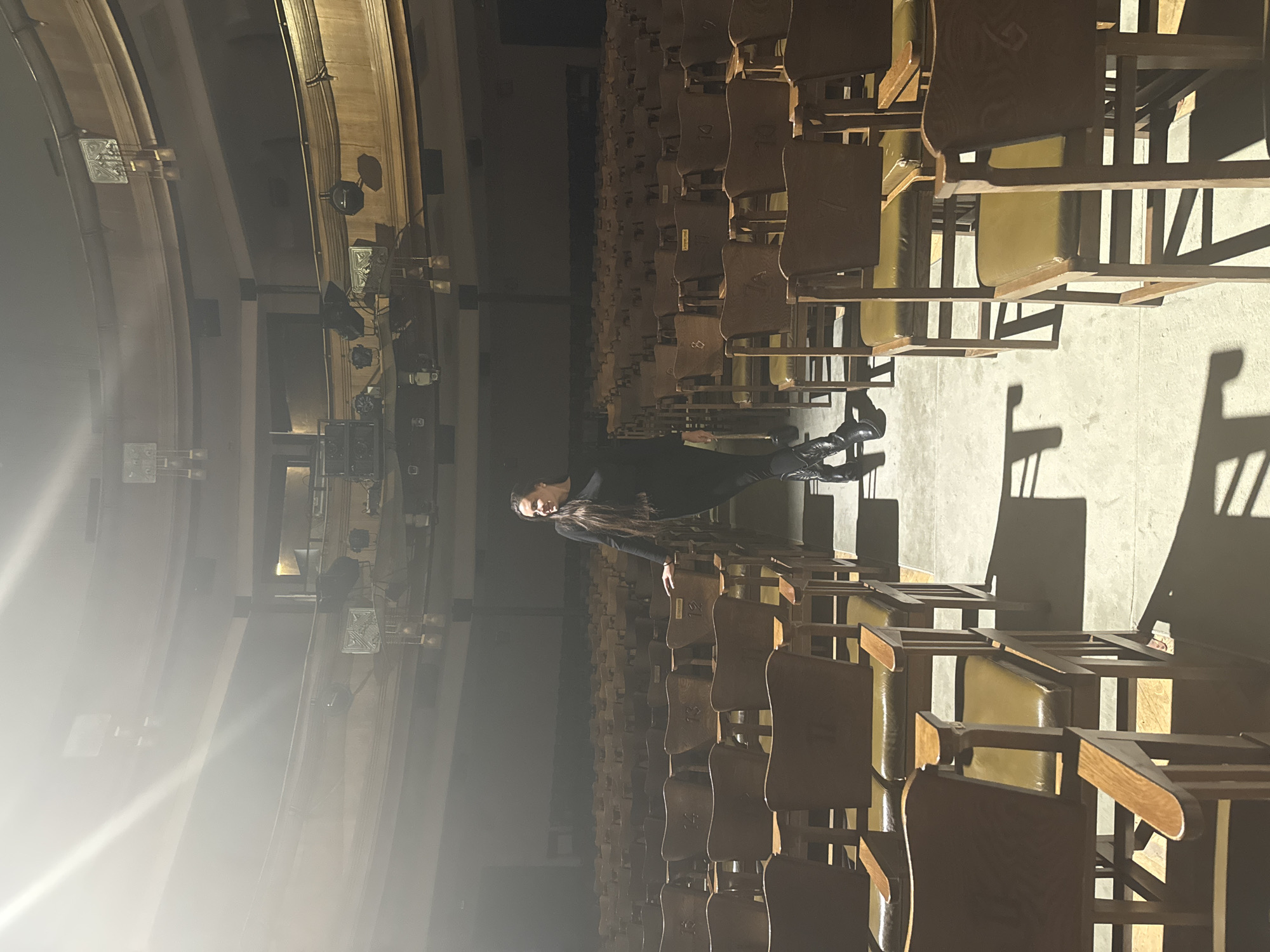 An eye-level, horizontally oriented photograph shows a woman standing in an auditorium with many rows of chairs. The scene may be Moscow, Russia. The rows of chairs appear to be aligned, stretching into the distance. The woman is standing in the aisle between the rows. She has long hair. She is wearing dark clothing. The auditorium has an ornate design. There is a bright light source on the left. The walls have a curved design, and there appear to be light fixtures on the walls. The room appears to be dimly lit, except for the bright light source. The chairs cast shadows on the floor.