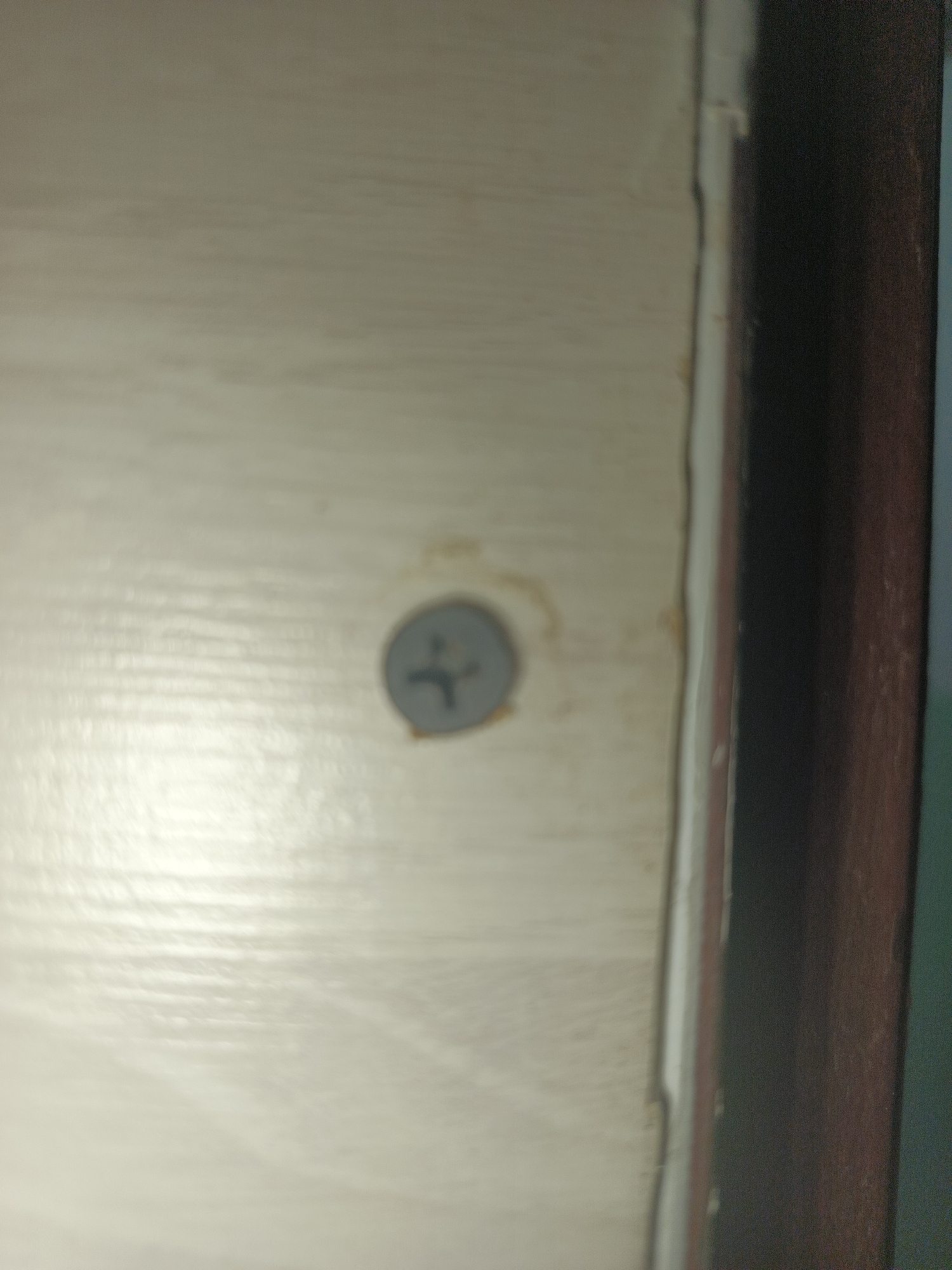 The image shows a close-up of a light-colored wooden surface, possibly a door or furniture, with a single screw embedded in it. The screw head is gray and has a cross-shaped indentation. There are some faint yellowish marks on the surface around the screw. To the right of the wooden surface, there is a vertical brown object, likely a door frame or trim, and a darker vertical area, possibly a shadow or another surface. The overall lighting suggests an indoor environment. The image does not contain any recognizable landmarks or contextual information that would allow for the identification of its location in Smolensk, Russia.