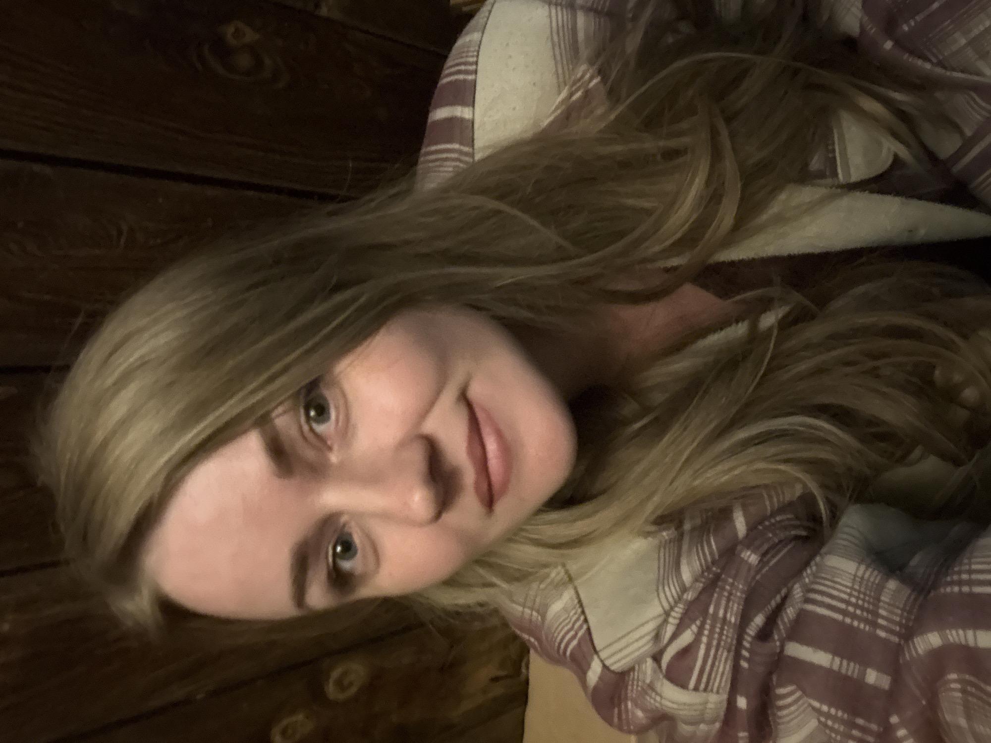 A selfie shows a woman with long, wavy blonde hair, wearing a plaid shirt. The background appears to be wooden wall paneling. There is no information in the image to confirm that it was taken in Chaikovsky, Russia.
