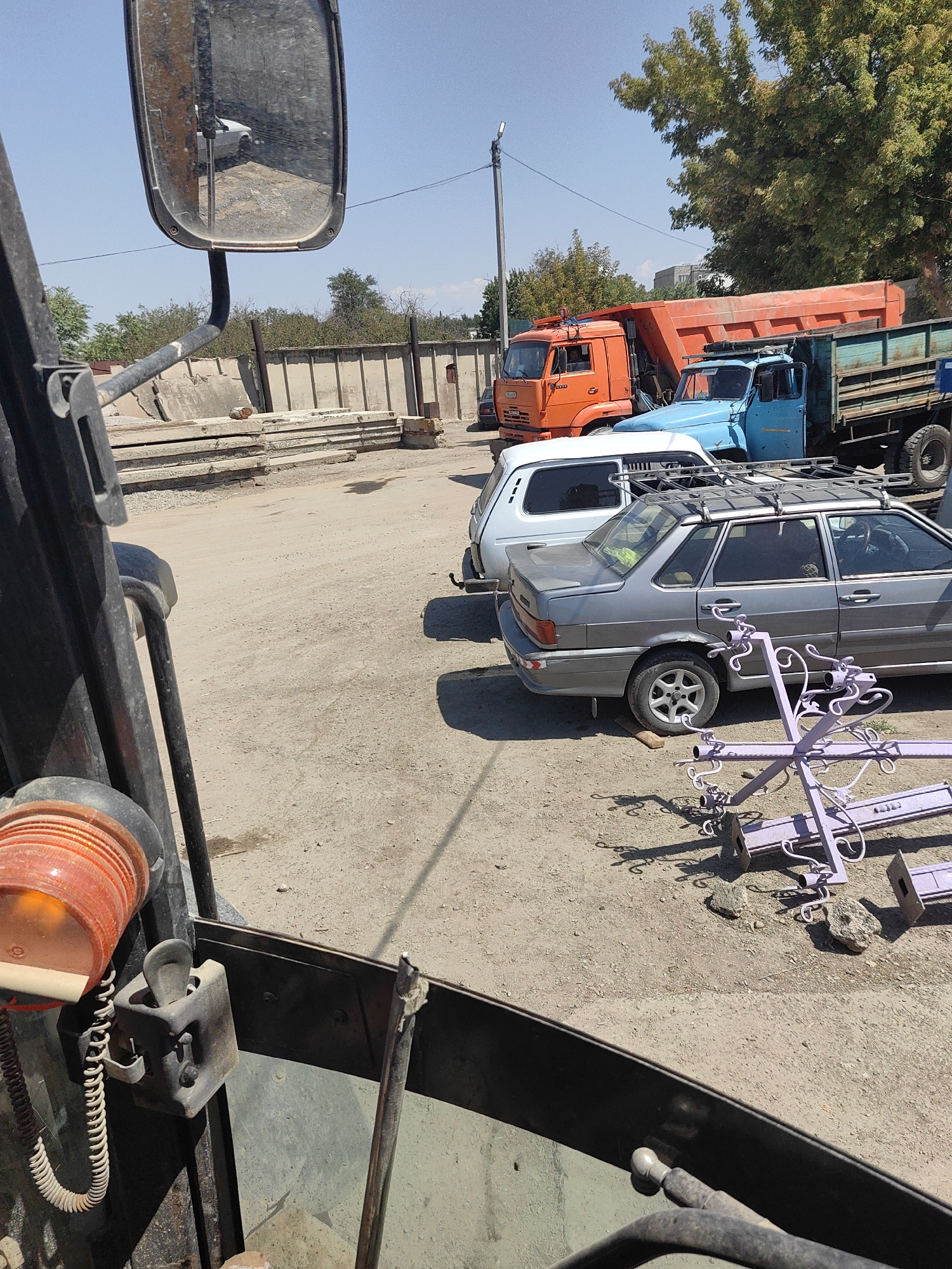 The image depicts an outdoor scene in Shymkent, Kazakhstan, likely a construction or industrial area. In the foreground, partially visible, is the interior of a vehicle, possibly a loader or truck, with an orange flashing light and a black control panel. Dominating the mid-ground are several vehicles: a silver sedan, a light blue hatchback, and two large trucks, one orange dump truck and another with a teal cargo bed. The ground is unpaved, with dirt and gravel. In the background, a concrete wall and stacked concrete slabs suggest construction activity. Trees with green foliage are visible, and the sky is clear and blue. A decorative purple metal structure, possibly a fence or garden ornament, lies on the ground in the right mid-ground, casting shadows. The overall impression is one of a working environment under a bright, sunny sky.