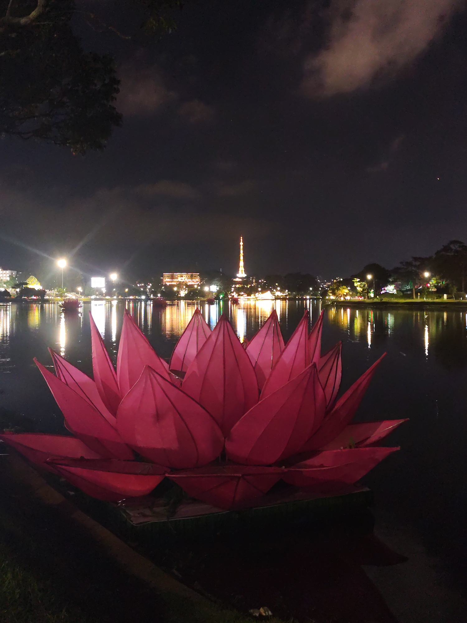 The image depicts a large, illuminated, pink lotus flower sculpture floating on a lake at night. The sculpture is positioned in the foreground. In the background, the city of Da Lat, Vietnam is visible across the water, identifiable by a tower structure with illuminated lights. Other buildings and streetlights are reflected in the dark water. The sky is dark with scattered clouds. Grass is visible in the lower left corner of the frame, suggesting a shoreline perspective.