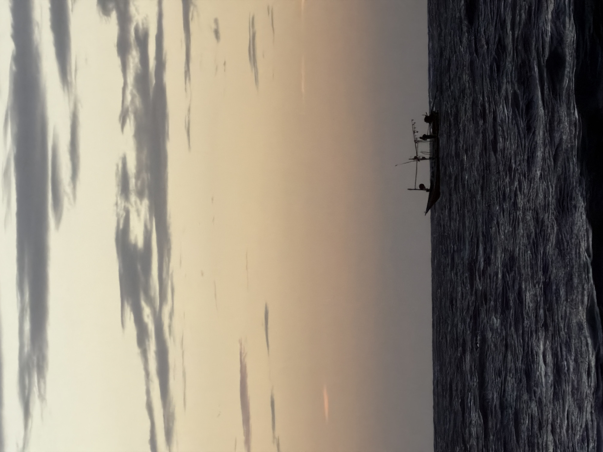 A fishing boat with two people aboard is silhouetted against a pastel-colored sky with scattered clouds. The boat is on dark, choppy water. The scene appears to be a coastal area in the Ko Samui District of Thailand.