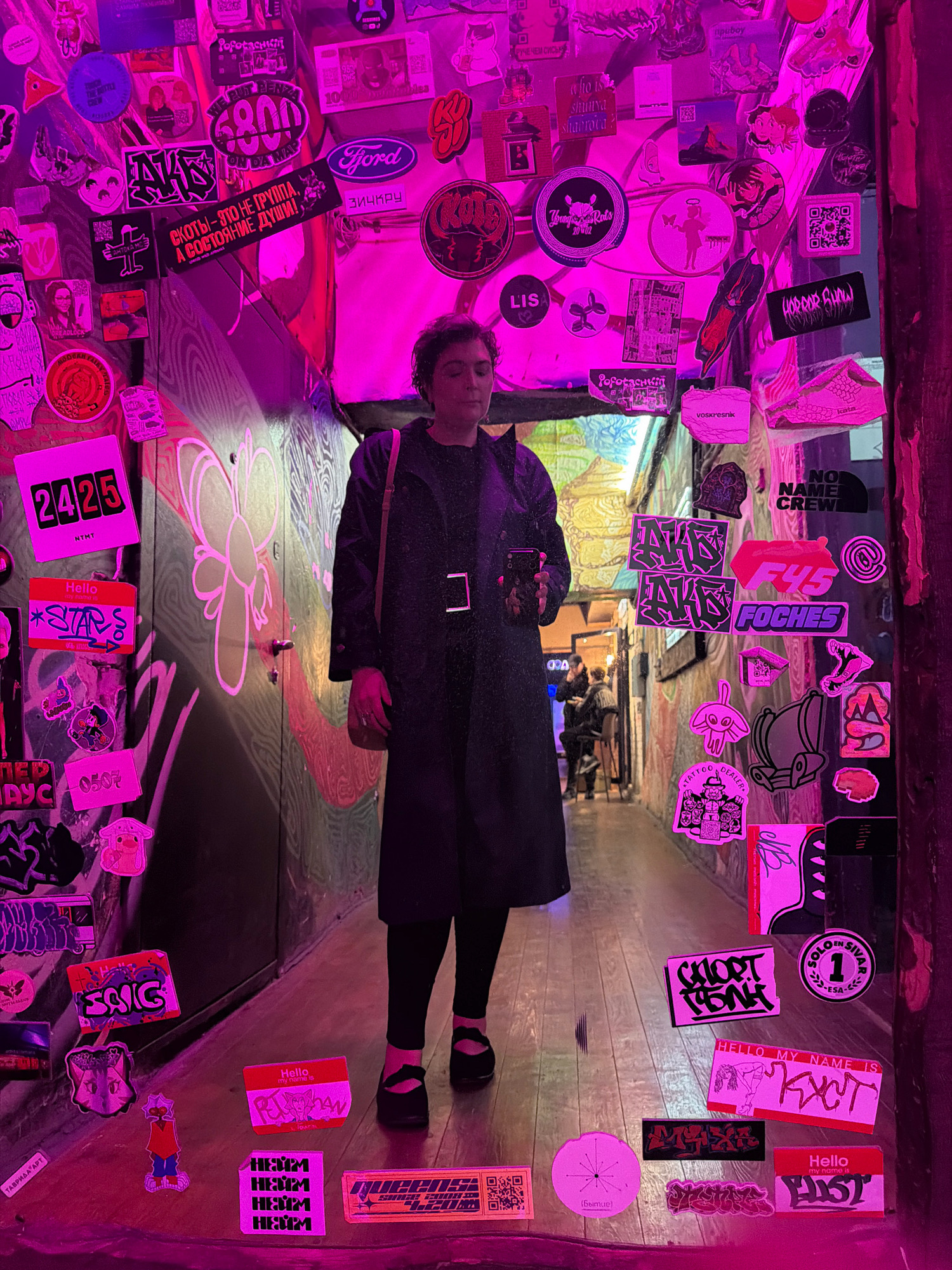 The image captures a dimly lit interior space, possibly a narrow alleyway or corridor, adorned with an abundance of stickers and graffiti. The walls are heavily decorated with various stickers, some appearing to be band merchandise, logos, and personal artwork. The color palette is dominated by purples and pinks, likely from ambient lighting.
In the center of the frame, a person with short, dark hair stands facing the camera. They are wearing a dark, long coat, black leggings, and black shoes with straps. The individual is holding a smartphone in their right hand. Their gaze is directed downwards, suggesting they are looking at the phone or the floor.
The environment appears to be a commercial or entertainment venue, given the presence of stickers, the graffiti, and the visible entryway in the background with people and furniture. The overall atmosphere is vibrant and urban, with a strong emphasis on street art and youth culture.
While the image is rich in visual details, specific indicators of its location within Samara, Russia, are not present. The stickers and graffiti are in Russian, indicating a Russian-speaking environment, but do not provide a precise geographical marker within the city.